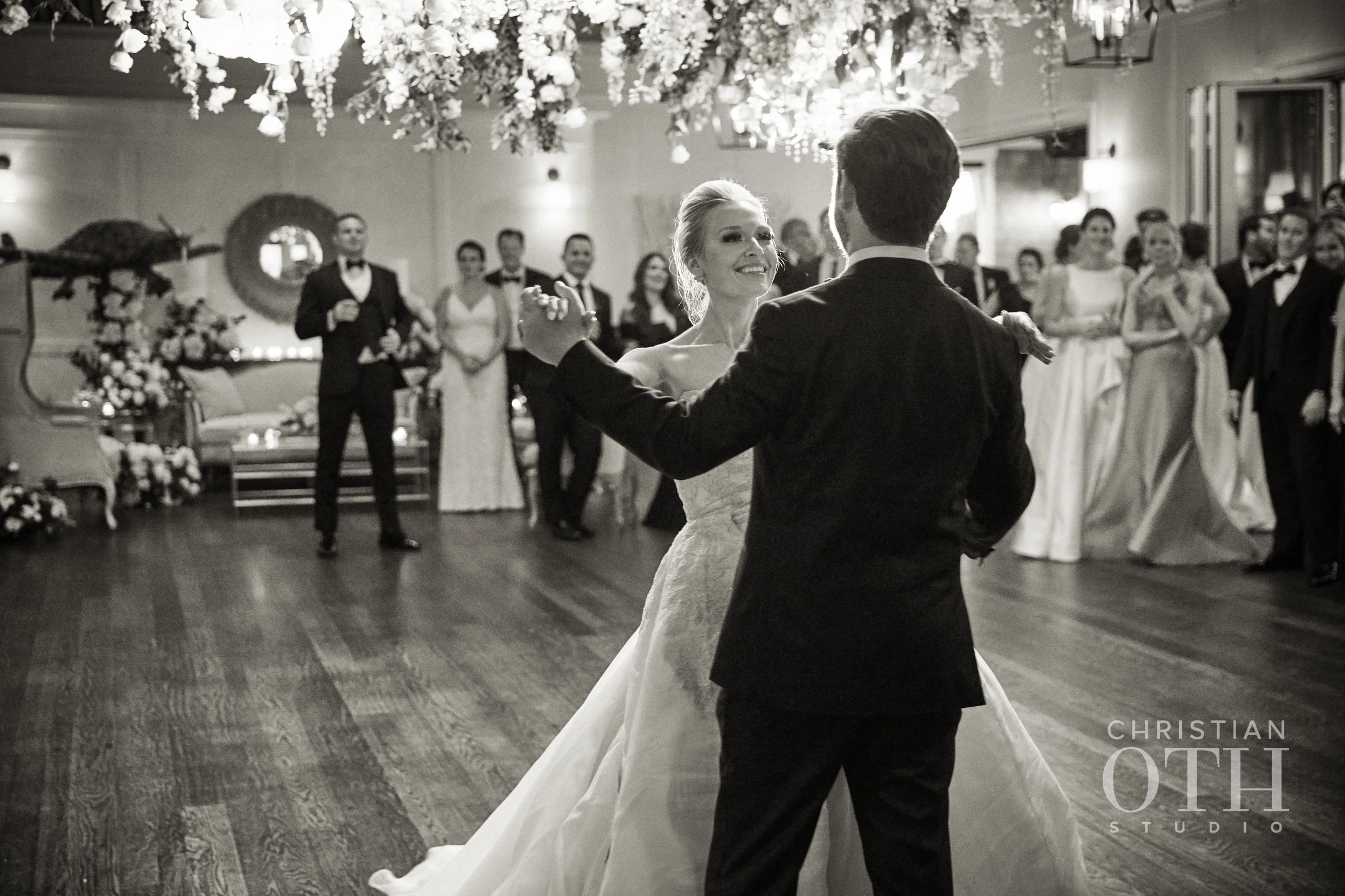 A bride and groom dance together in formal wedding attire on a polished wooden floor, surrounded by wedding guests in formal dresses and tuxedos, under hanging floral decorations.