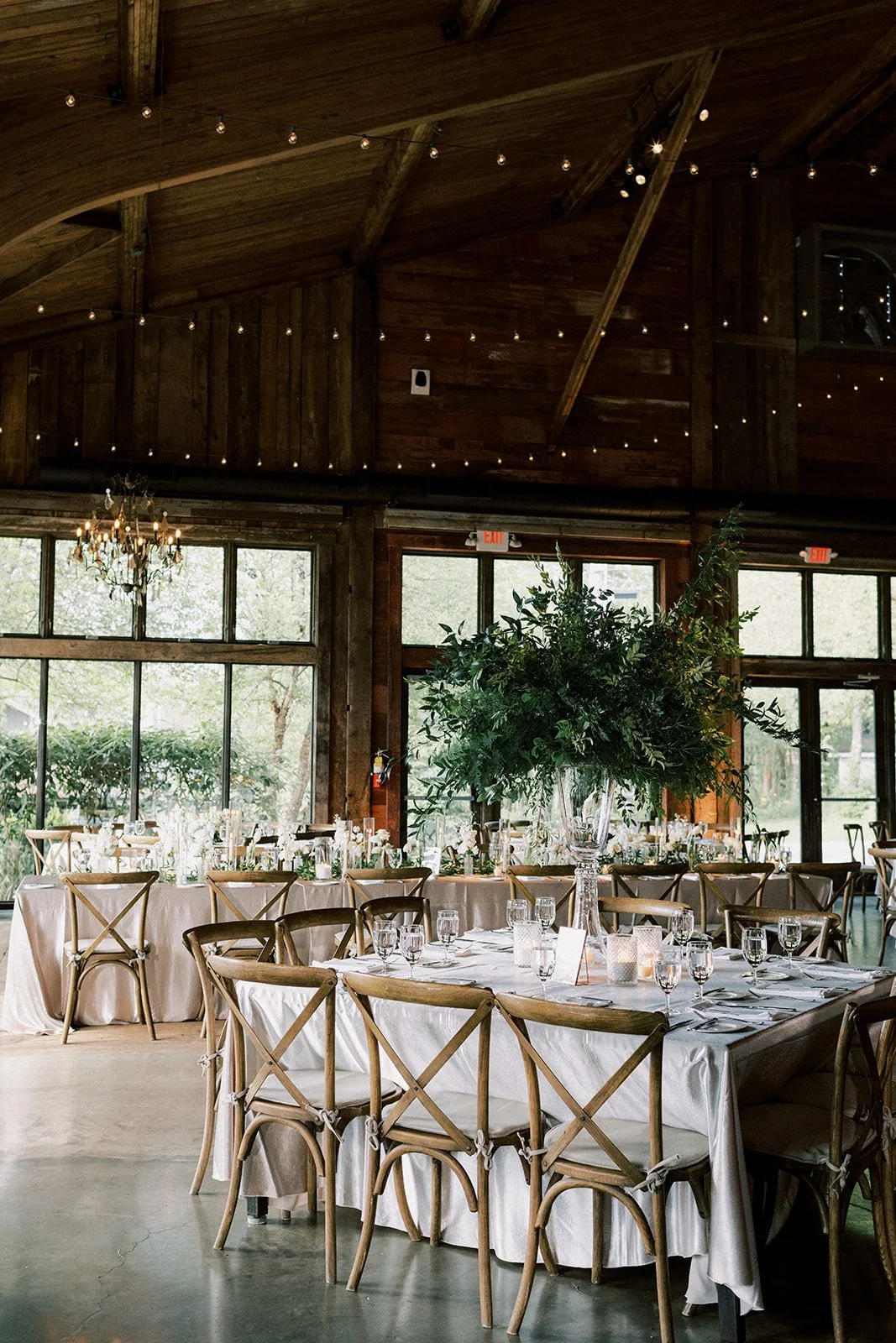 Elegant indoor wedding reception area with round tables, chairs, and a large floral centerpiece. The room features wooden walls, large windows, string lights, and a chandelier.