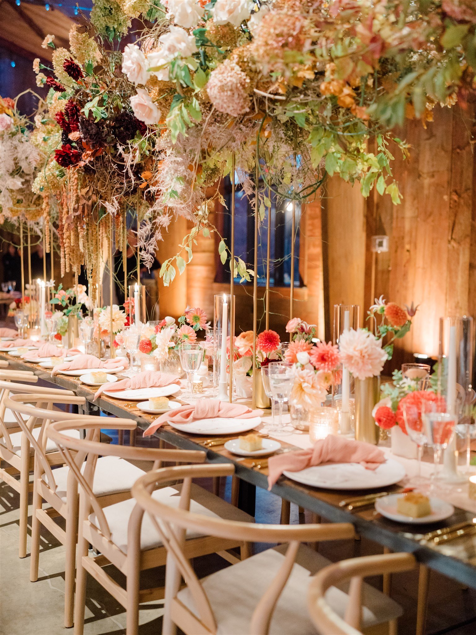 A beautifully decorated banquet table set with white plates, pink napkins, slices of cake, and elegant glassware. The table is adorned with tall candles and vibrant pink, peach, and white flowers in gold vases. Above, a large floral arrangement with 