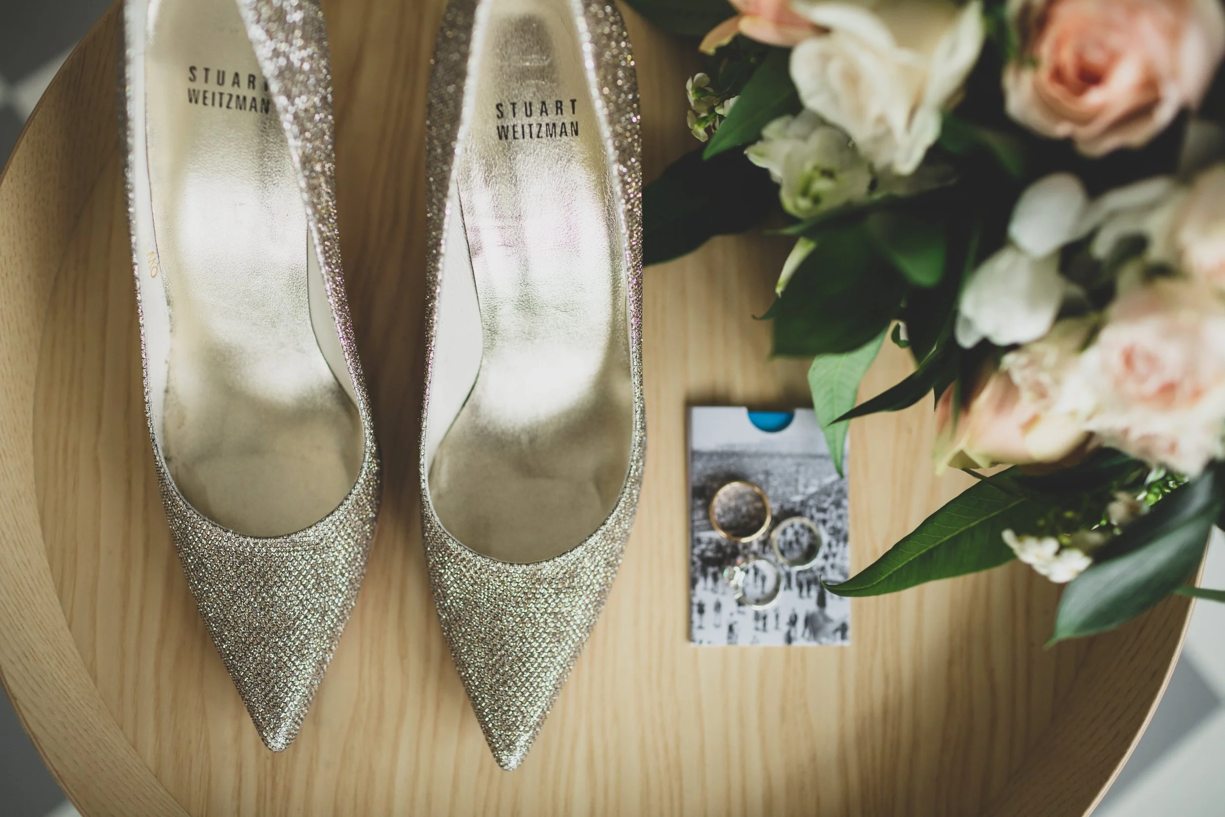 A pair of glittery, pointed-toe high heels, wedding rings on a card, and a bouquet of pink and white flowers on a wooden surface.