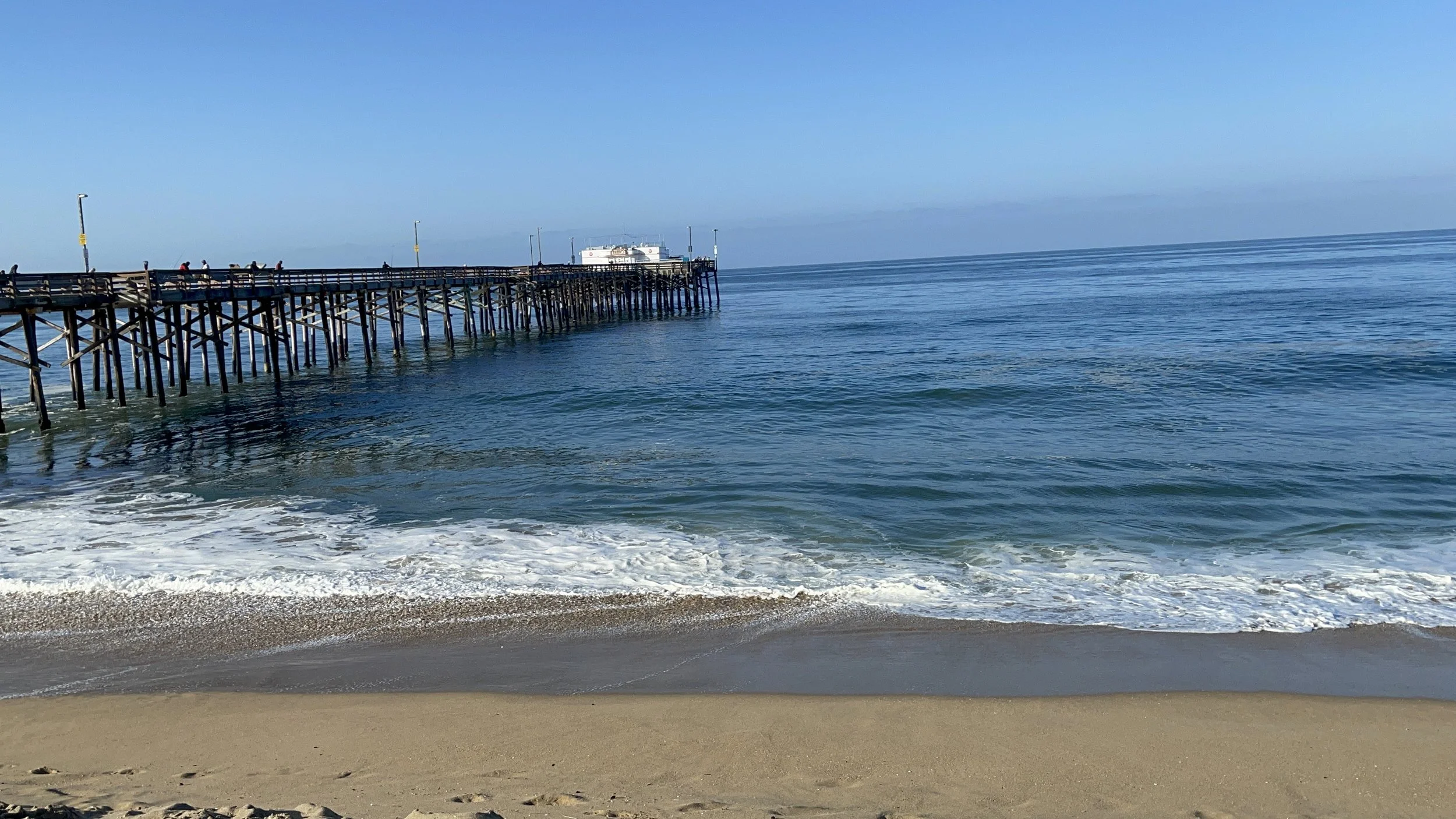 A wooden pier extending into the ocean with people walking on it, clear blue sky, calm waters, and sandy beach in the foreground.