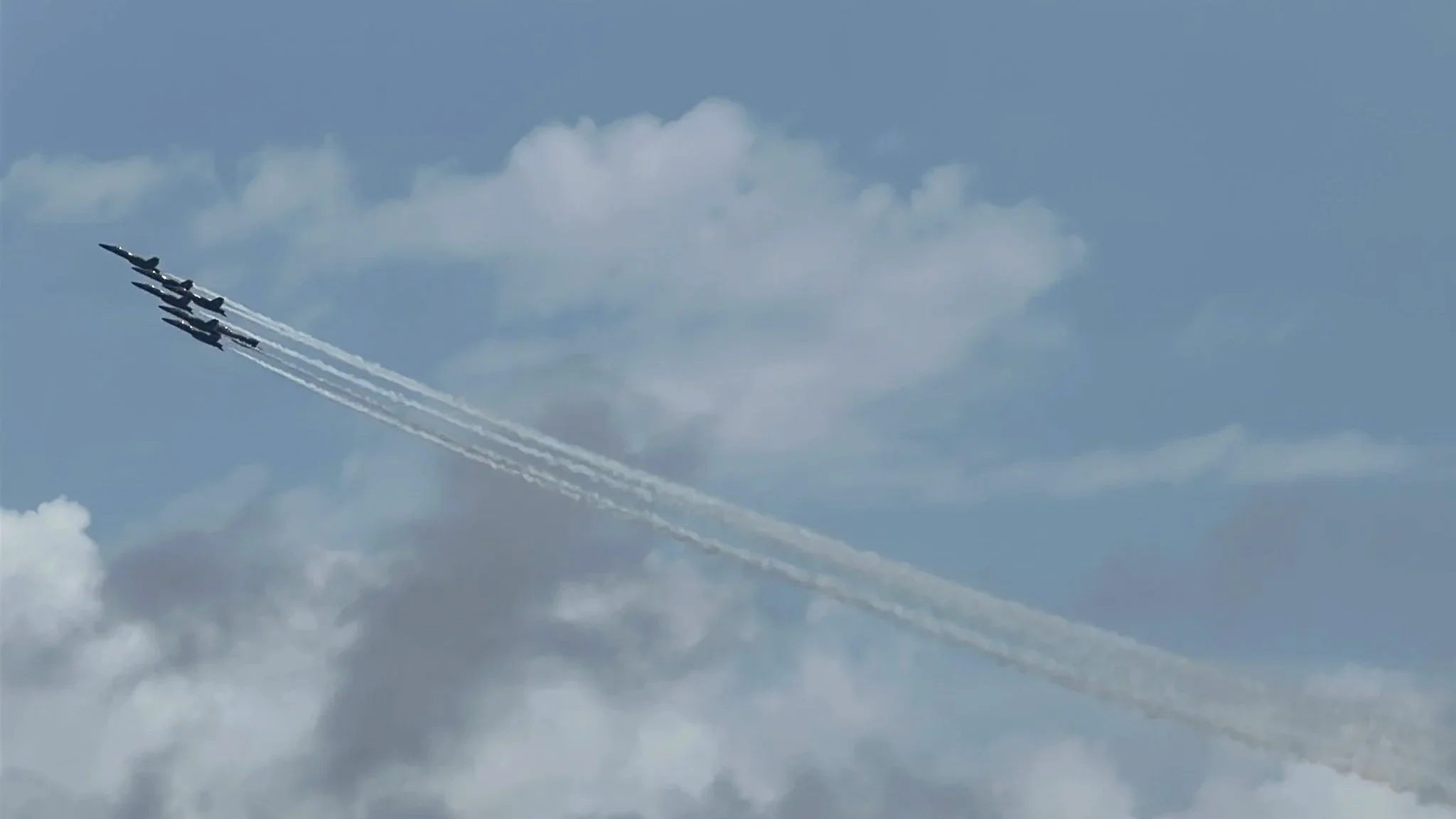 Six fighter jets flying in a tight formation, leaving white smoke trails across a partly cloudy sky.