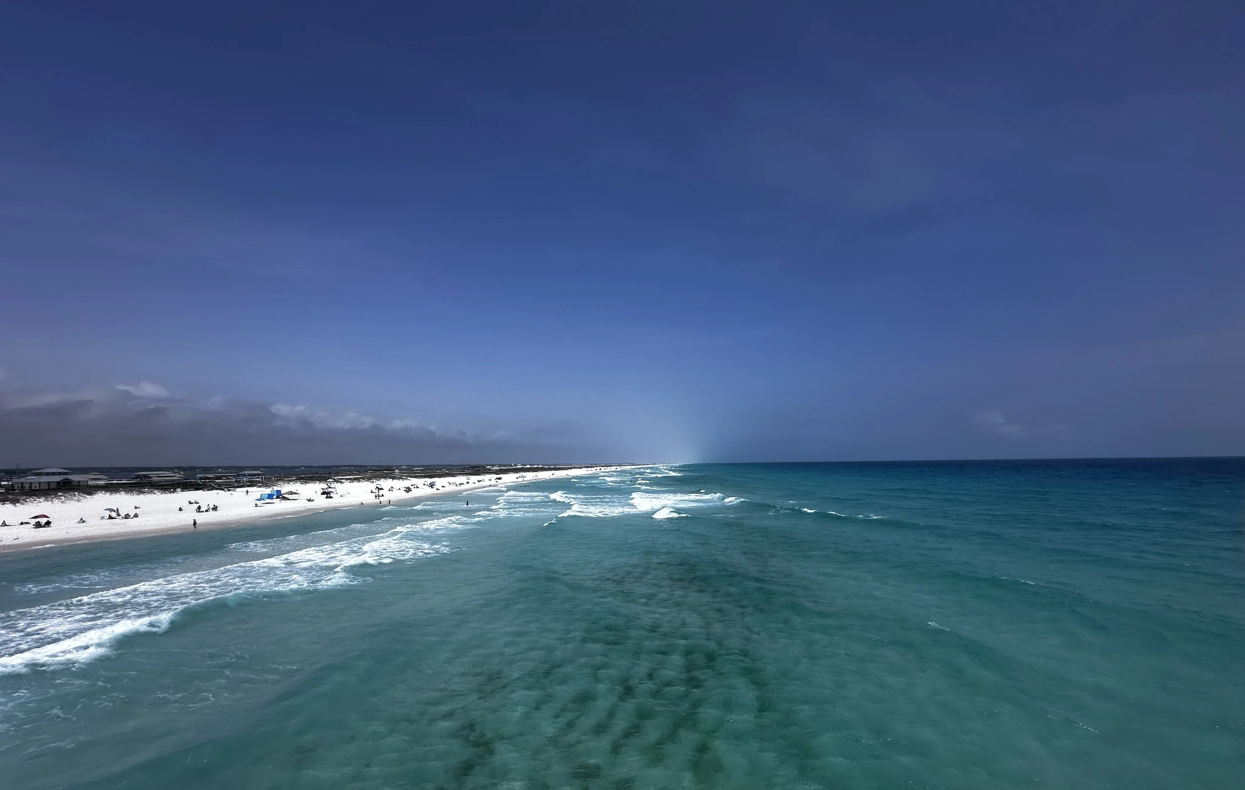 A beach scene with white sandy shoreline, turquoise ocean water, and people relaxing under umbrellas. The sky is clear with some clouds on the horizon.