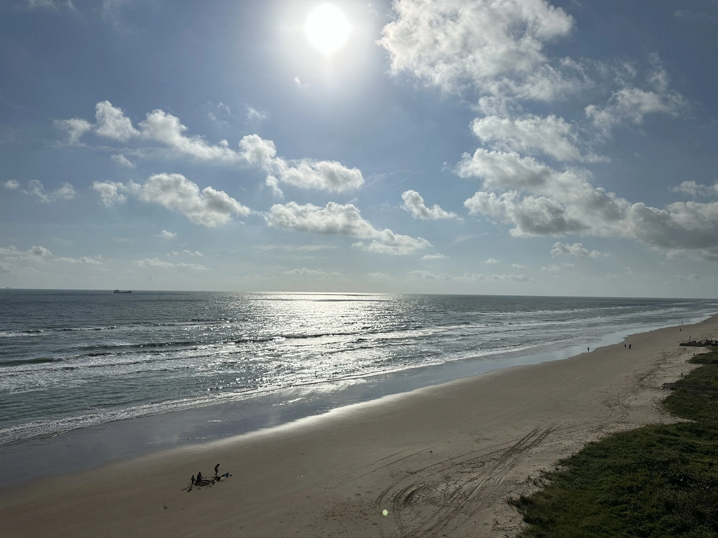 A beach scene with the sun high in the partly cloudy sky, reflecting sunlight on the ocean waves. There are a few people on the sandy shore near the water, and a ship can be seen in the distance on the horizon.