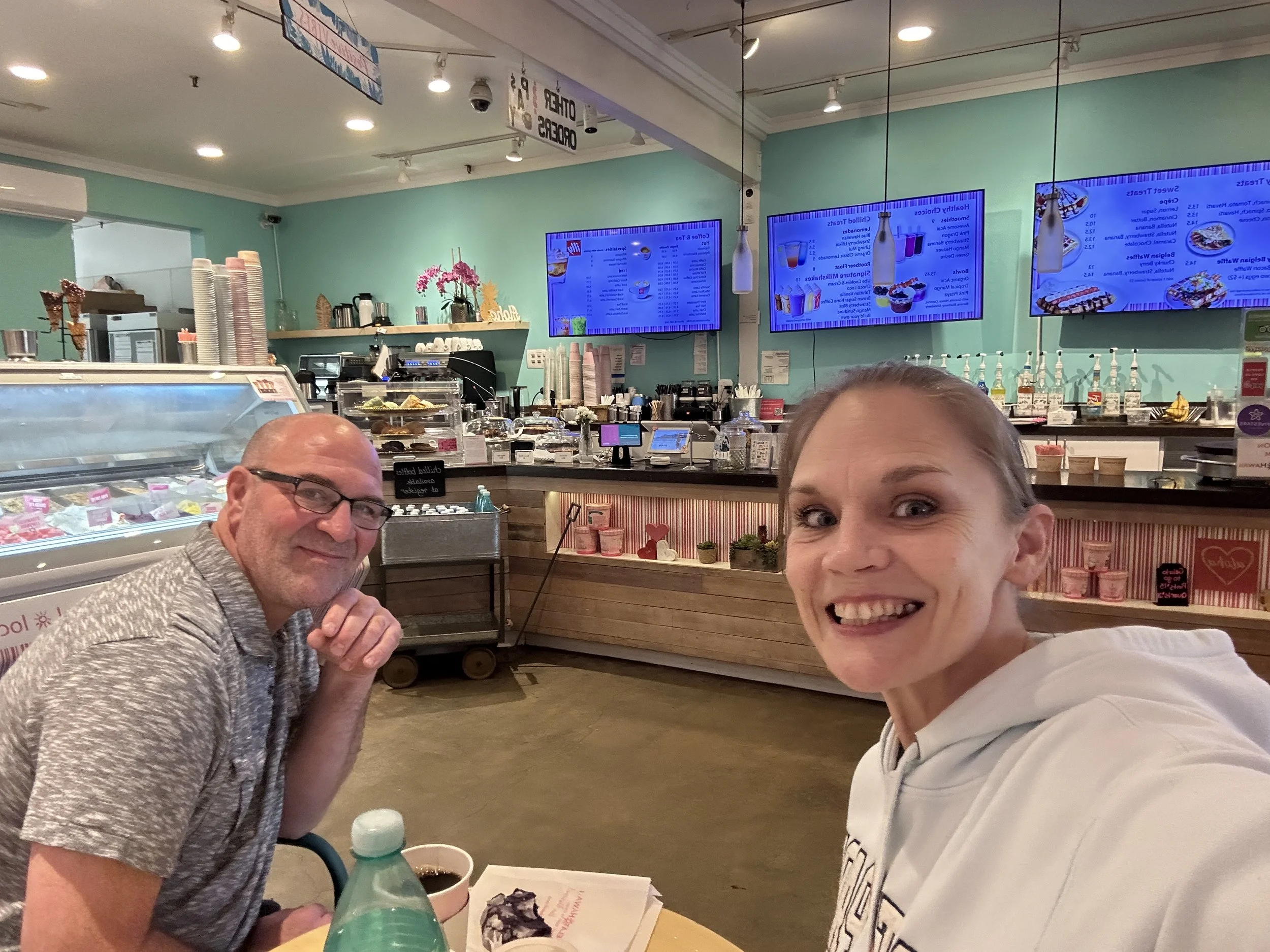 Two people sitting at a table inside a dessert shop, smiling at the camera. The man is wearing glasses and a patterned shirt, and the woman is wearing a white hoodie. The background features digital menu screens, stacked paper cups, and ice cream displays.