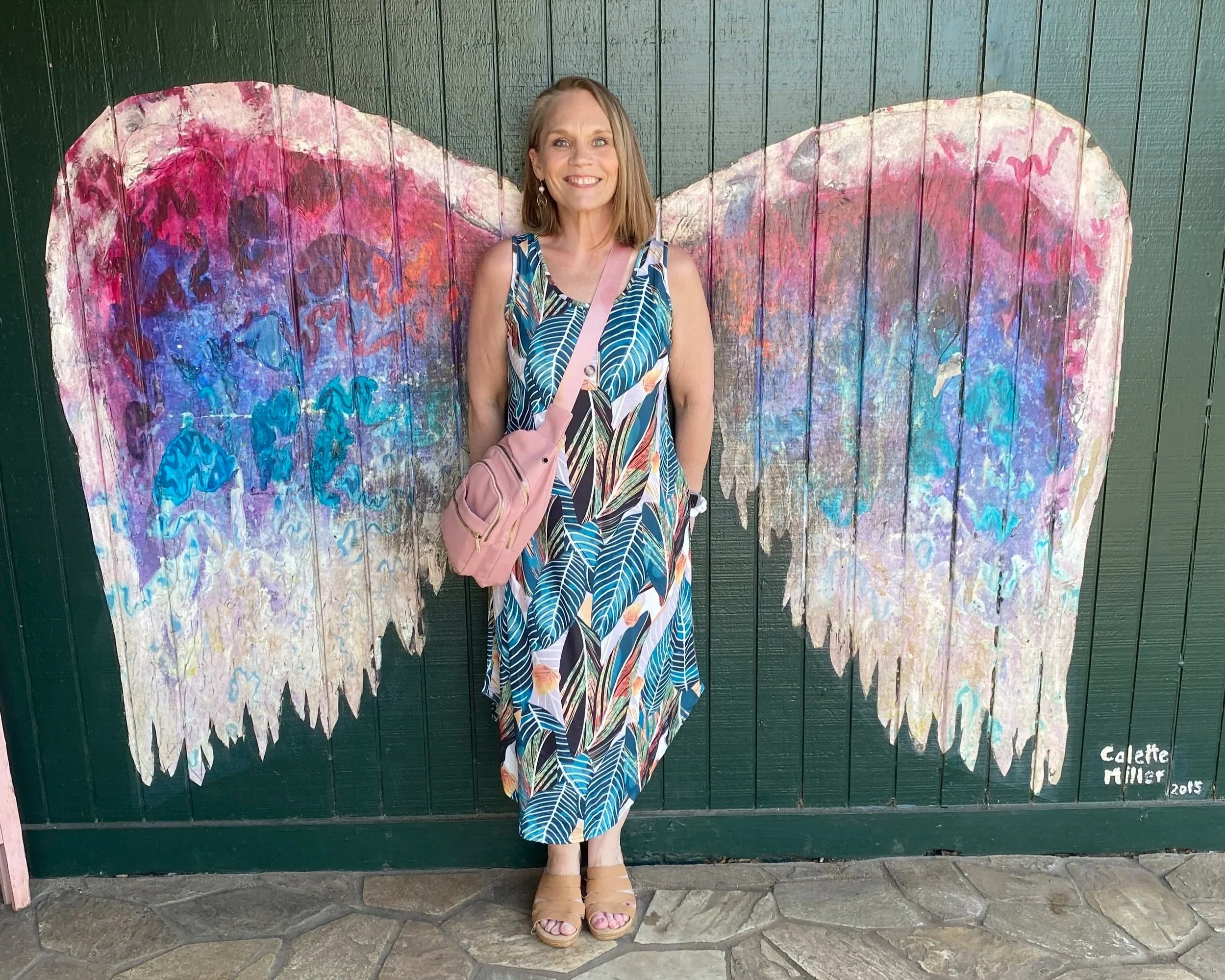 Woman in a tropical patterned dress standing in front of a heart-shaped painted mural on a green wooden wall.