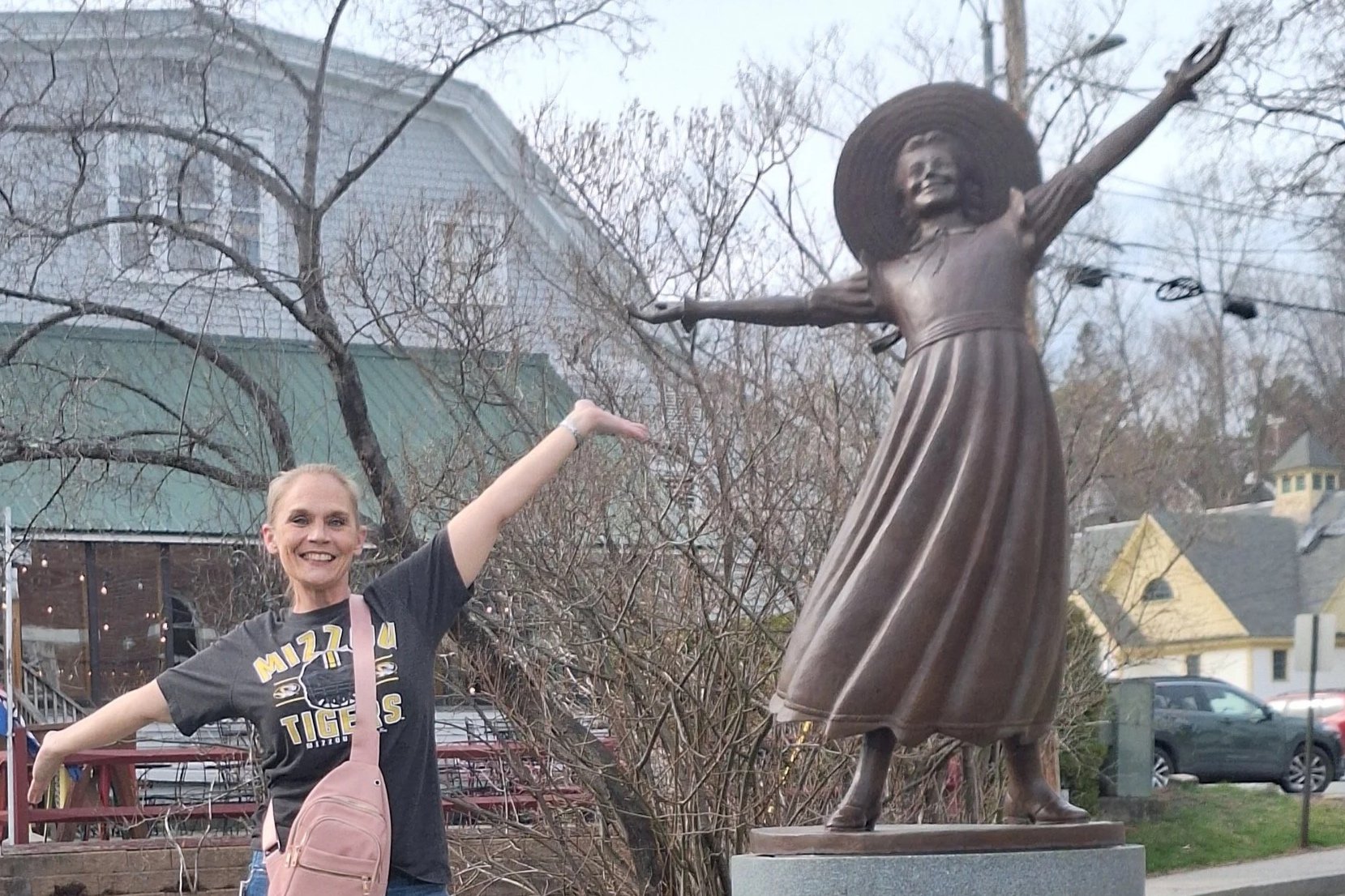 A woman smiling and posing with her arm extended toward a statue of a young girl in a dress with a wide-brimmed hat, standing on a platform in an outdoor setting with trees, houses, and parked cars in the background.