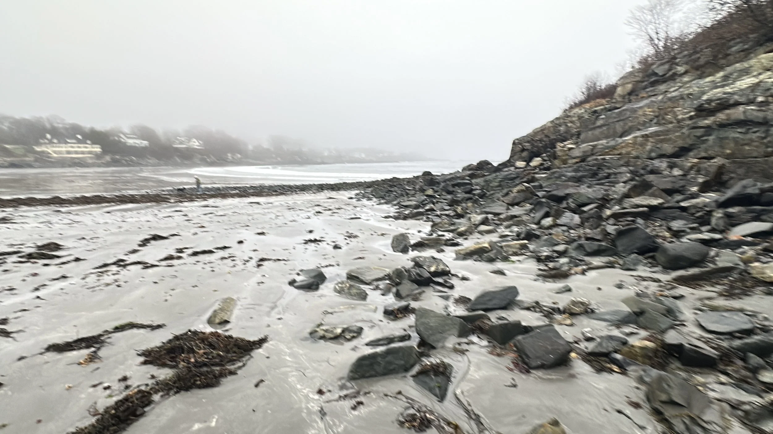 A rocky shoreline with a pebble-covered beach, a cliff on the right, and foggy weather in the background.