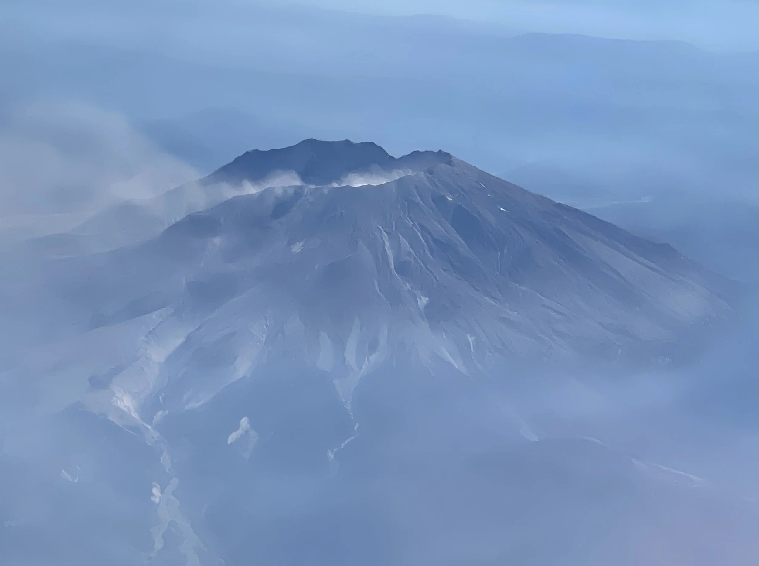 An aerial view of a volcano emitting smoke and ash, with a mountain range in the background, surrounded by clouds and mist.