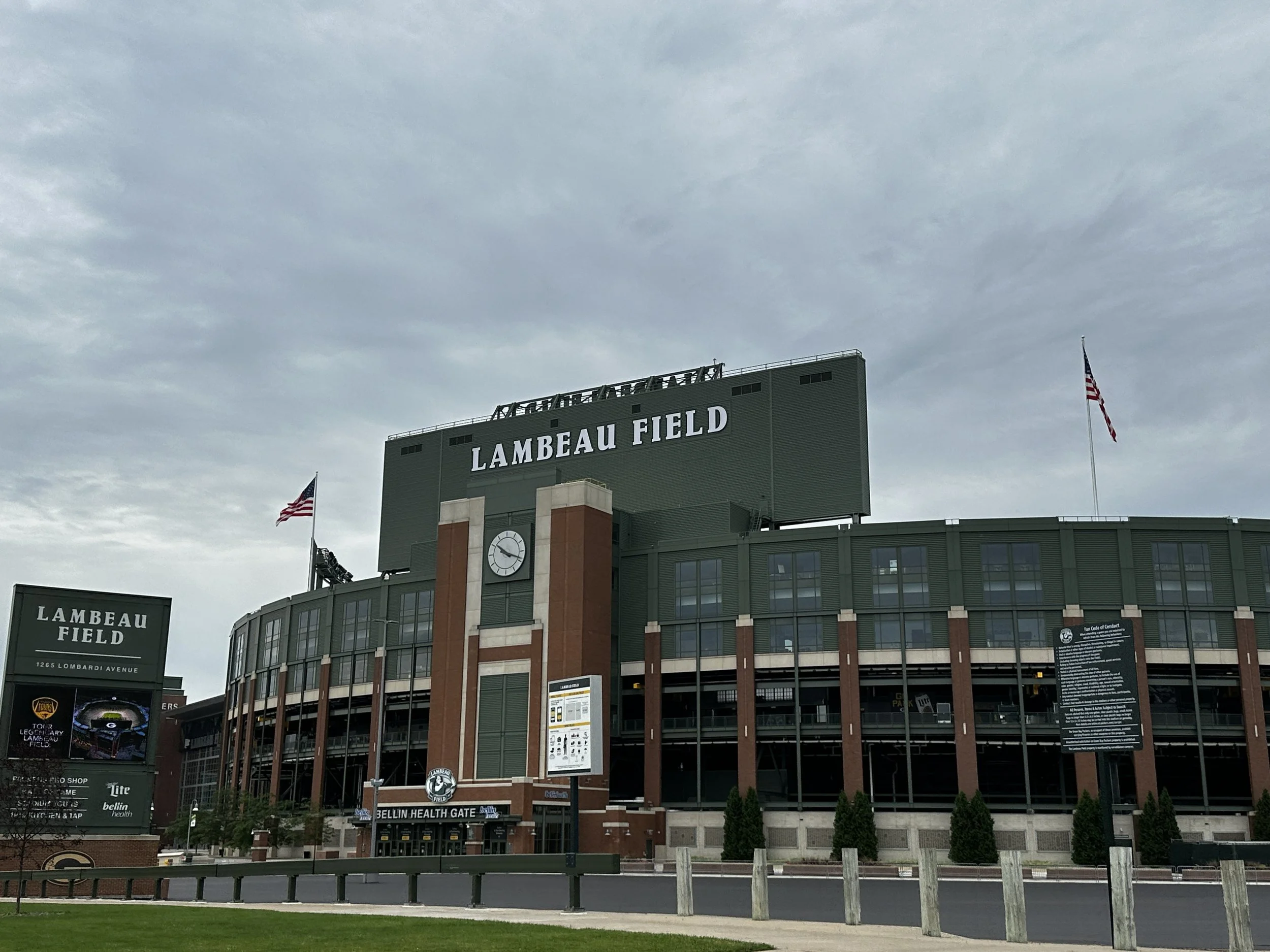 Exterior view of Lambeau Field stadium with overcast sky, American flags, and signage including Bellin Health Gate.