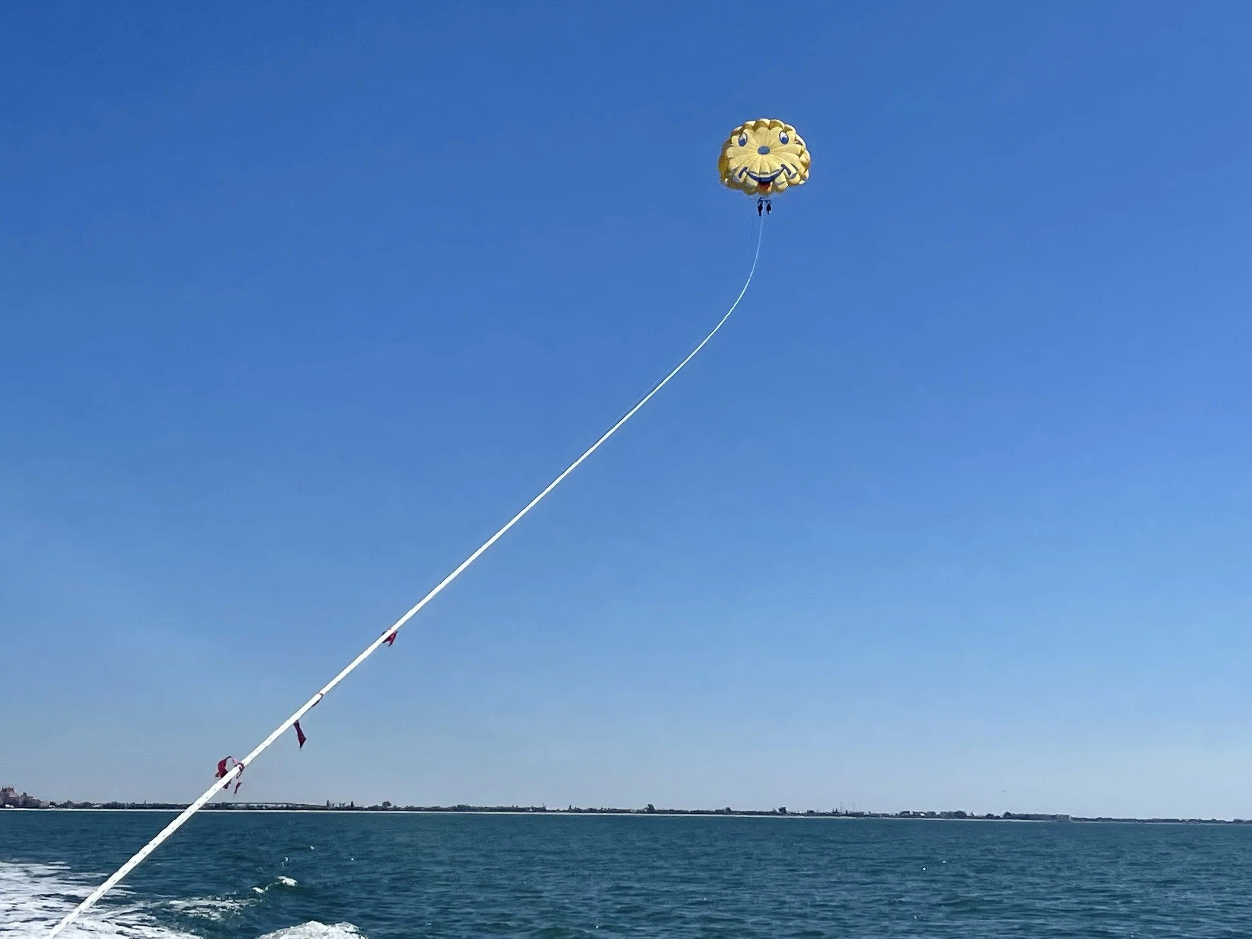 A yellow smiley face-shaped parachute or parasail floating in a clear blue sky, with a long white tow rope connected to a boat on the water below.