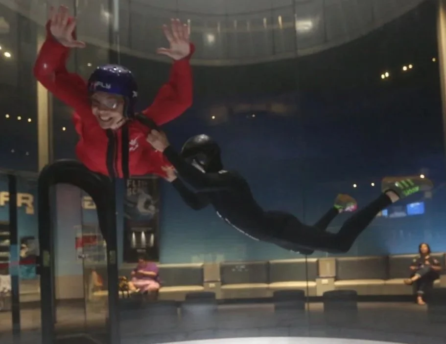 A person in a skydiving suit and helmet appears to be floating with the assistance of a wind tunnel, and another person in a red jacket and helmet is smiling and holding onto the first person in an indoor skydiving facility.