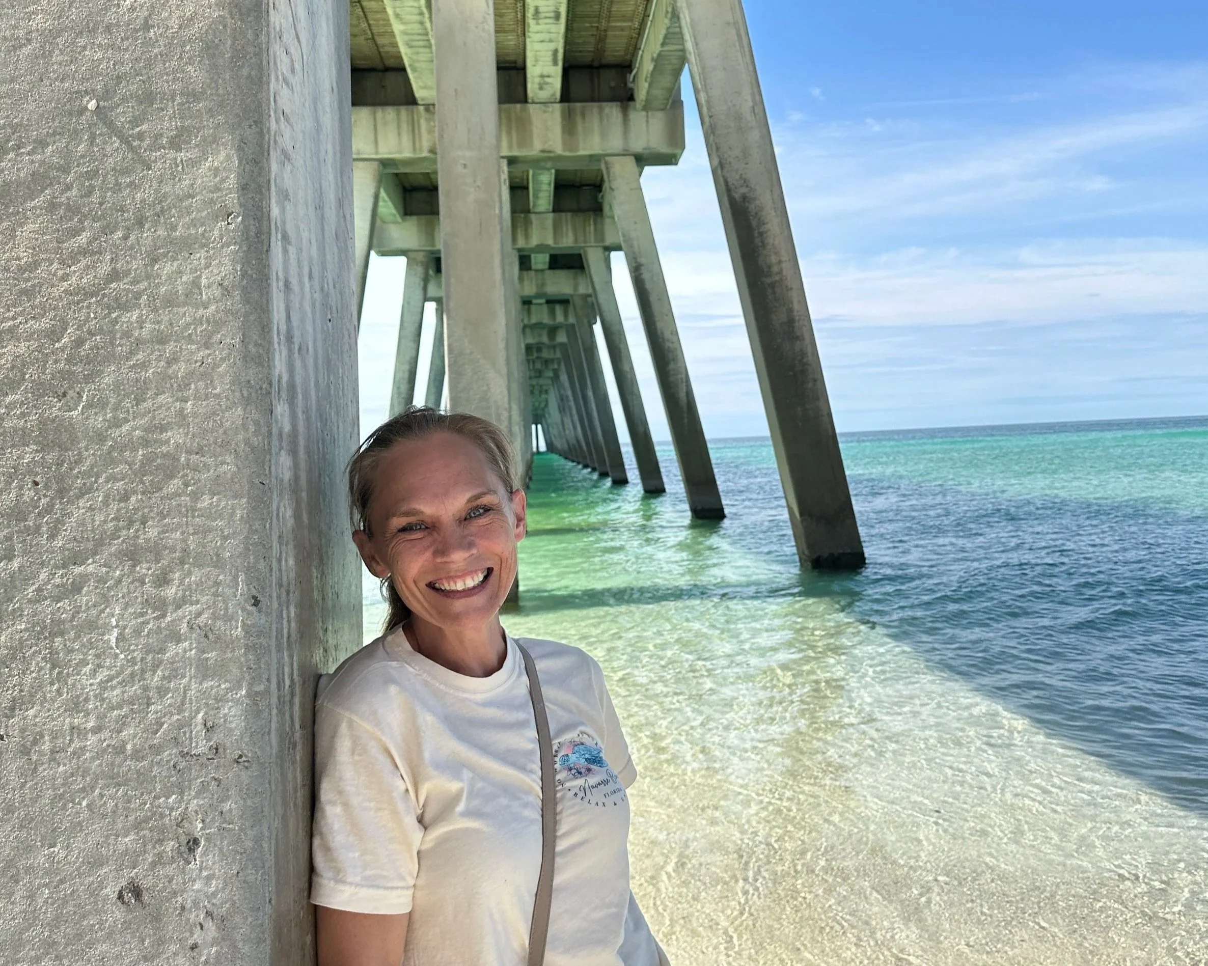 Smiling woman with blonde hair standing under a pier at the beach, with turquoise water and a blue sky in the background.