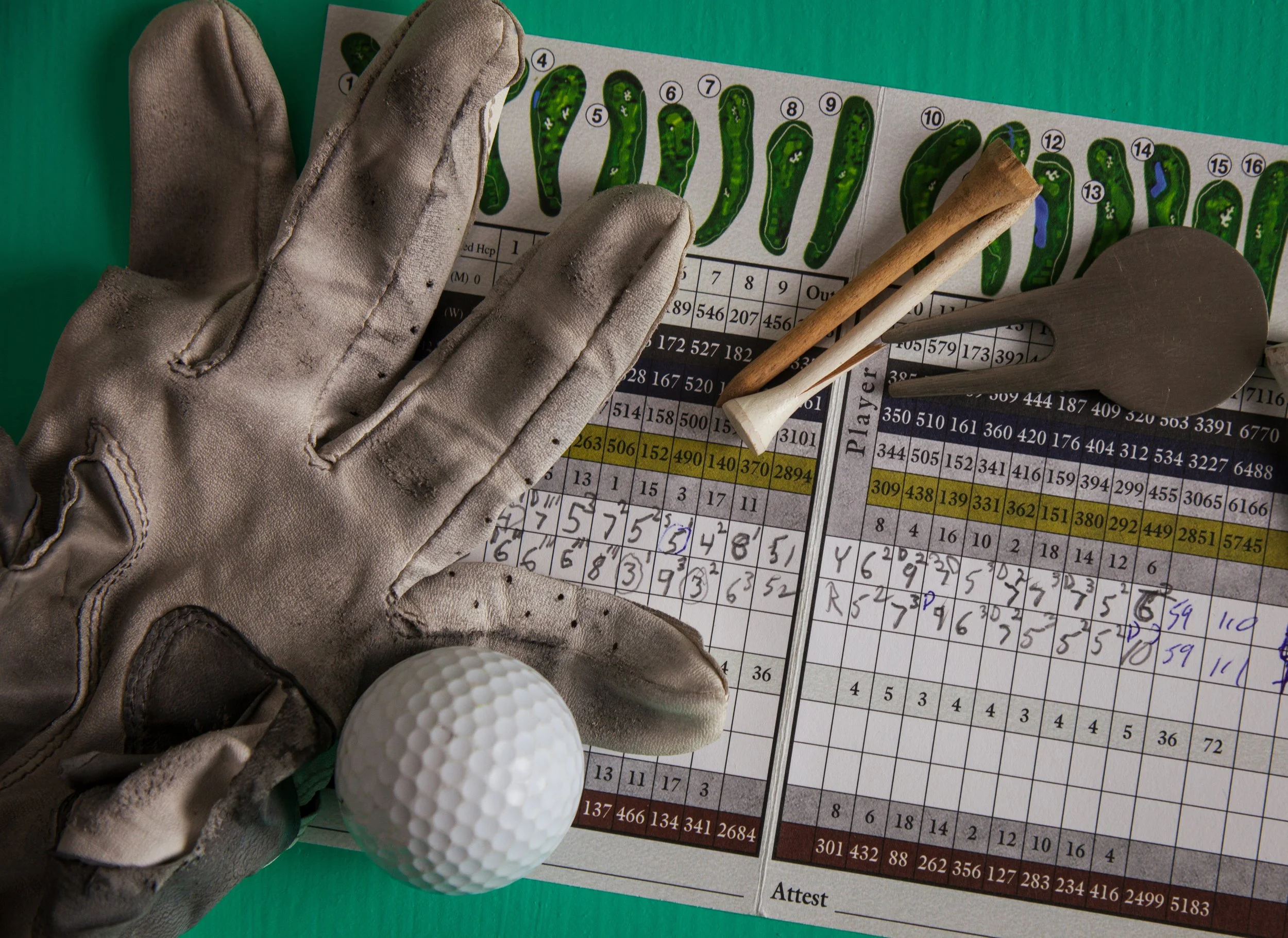 A close-up of a golf scorecard, two wooden golf tees, a golf glove, and a golf ball on a green background.