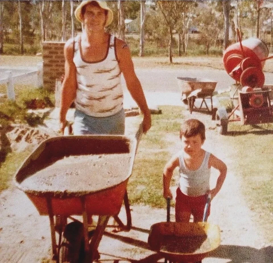 Woman and young boy outdoors, pushing wheelbarrows filled with dirt, with construction materials and a cement mixer in the background.