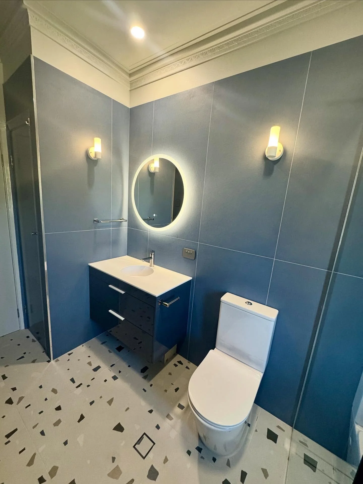 Modern bathroom with blue tiled walls, white toilet, vanity with a round mirror and illuminated border, wall sconces, and patterned tile floor.
