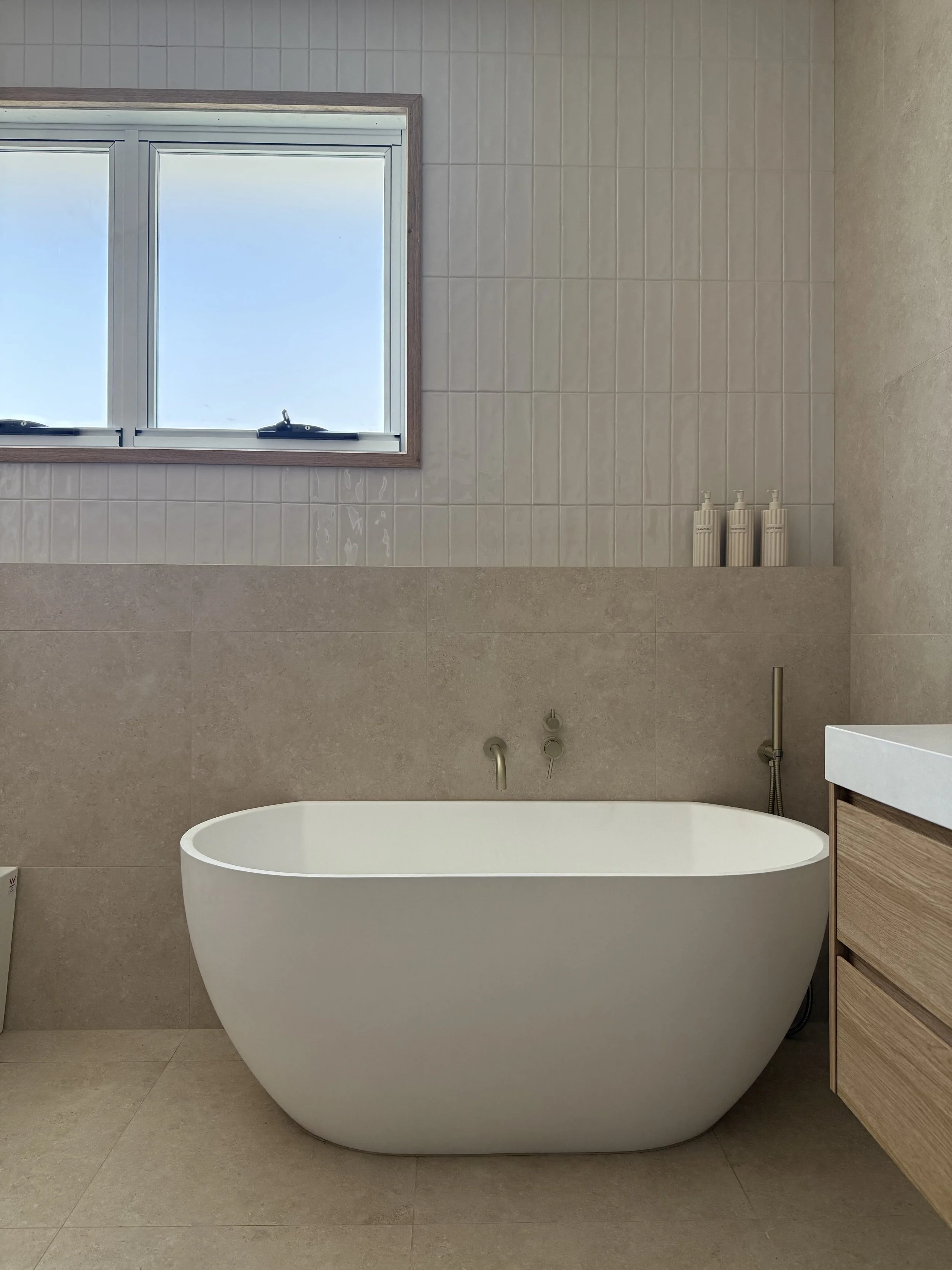 Modern bathroom with a freestanding white bathtub, a window with blue sky views, and beige tiled walls.