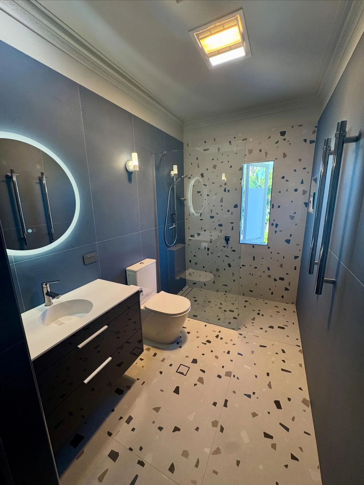 Modern bathroom with terrazzo tile floors and a terrazzo wall in the shower area, a round mirror with a light border above the sink, a white toilet, a glass shower enclosure with a window, and sleek cabinetry and fixtures.