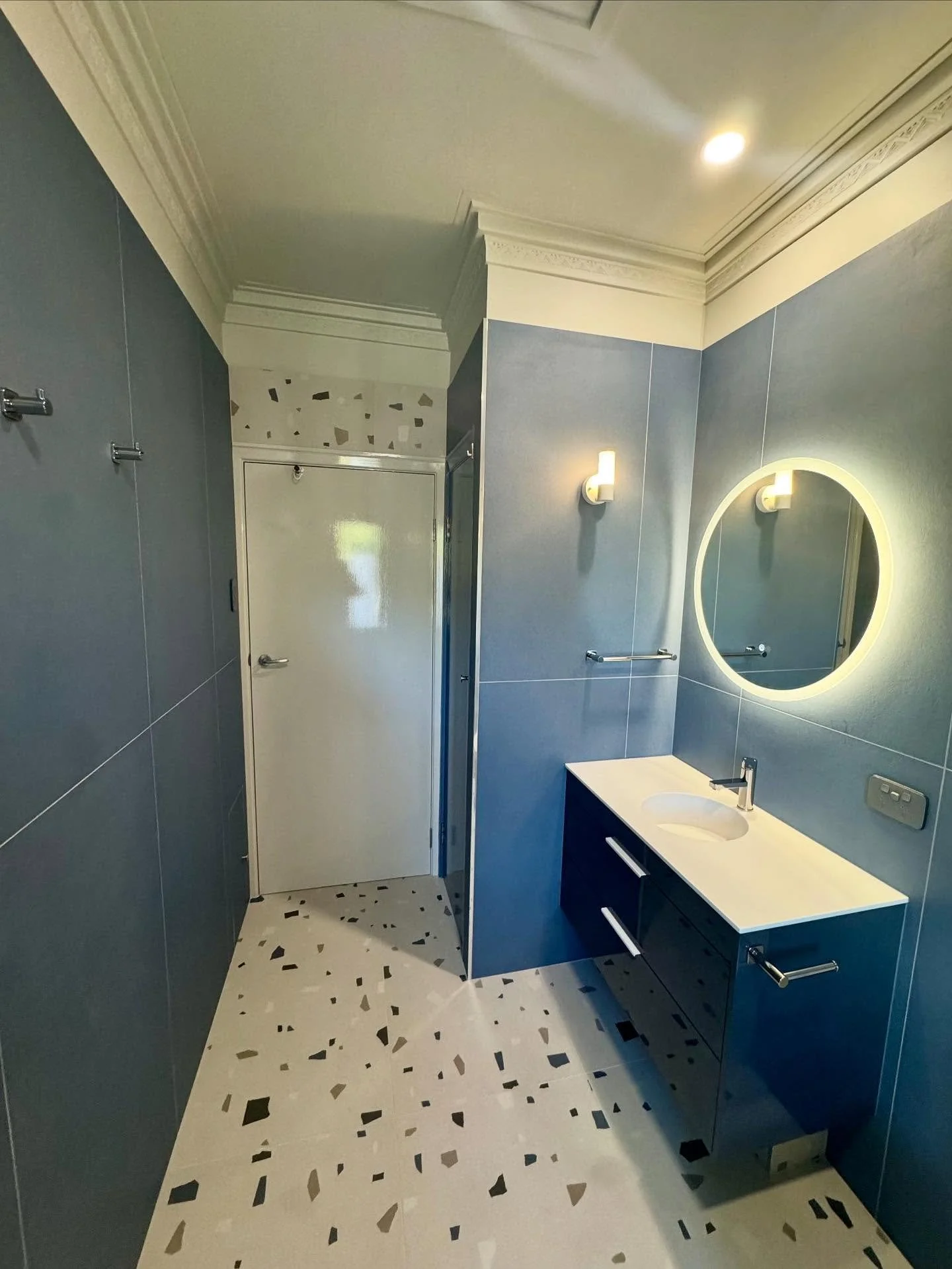 A modern bathroom with blue tiled walls and a terrazzo floor, featuring a round backlit mirror above a white countertop with a sink, wall-mounted light fixtures, and a door at the back.
