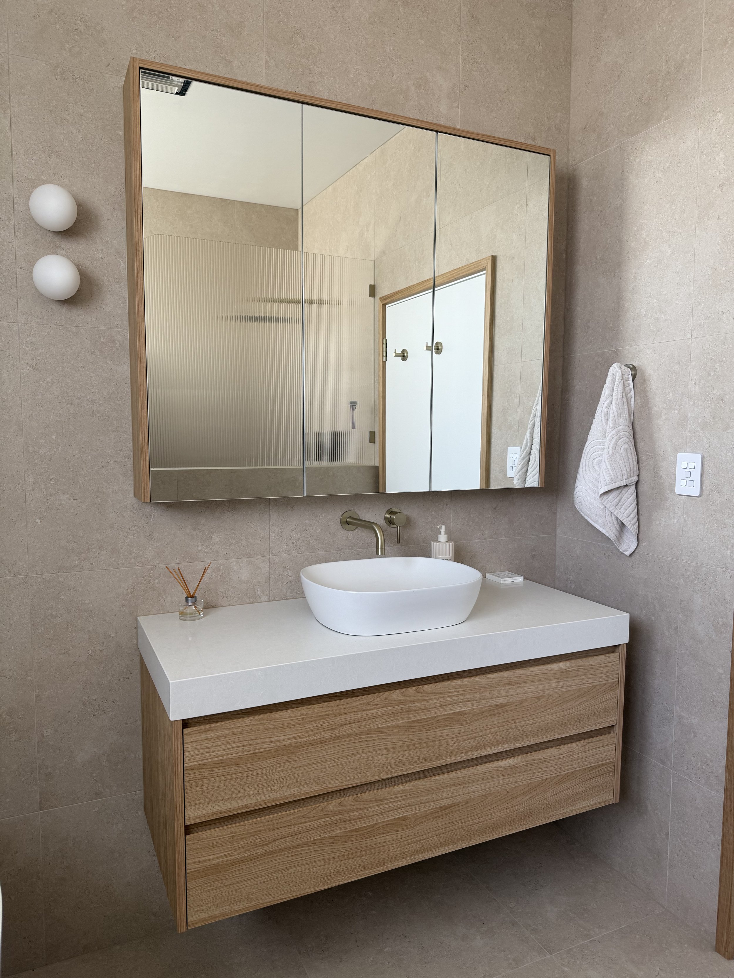 Modern bathroom with a wooden vanity, white countertop, oval vessel sink, large mirrored cabinet, and beige tiled walls.