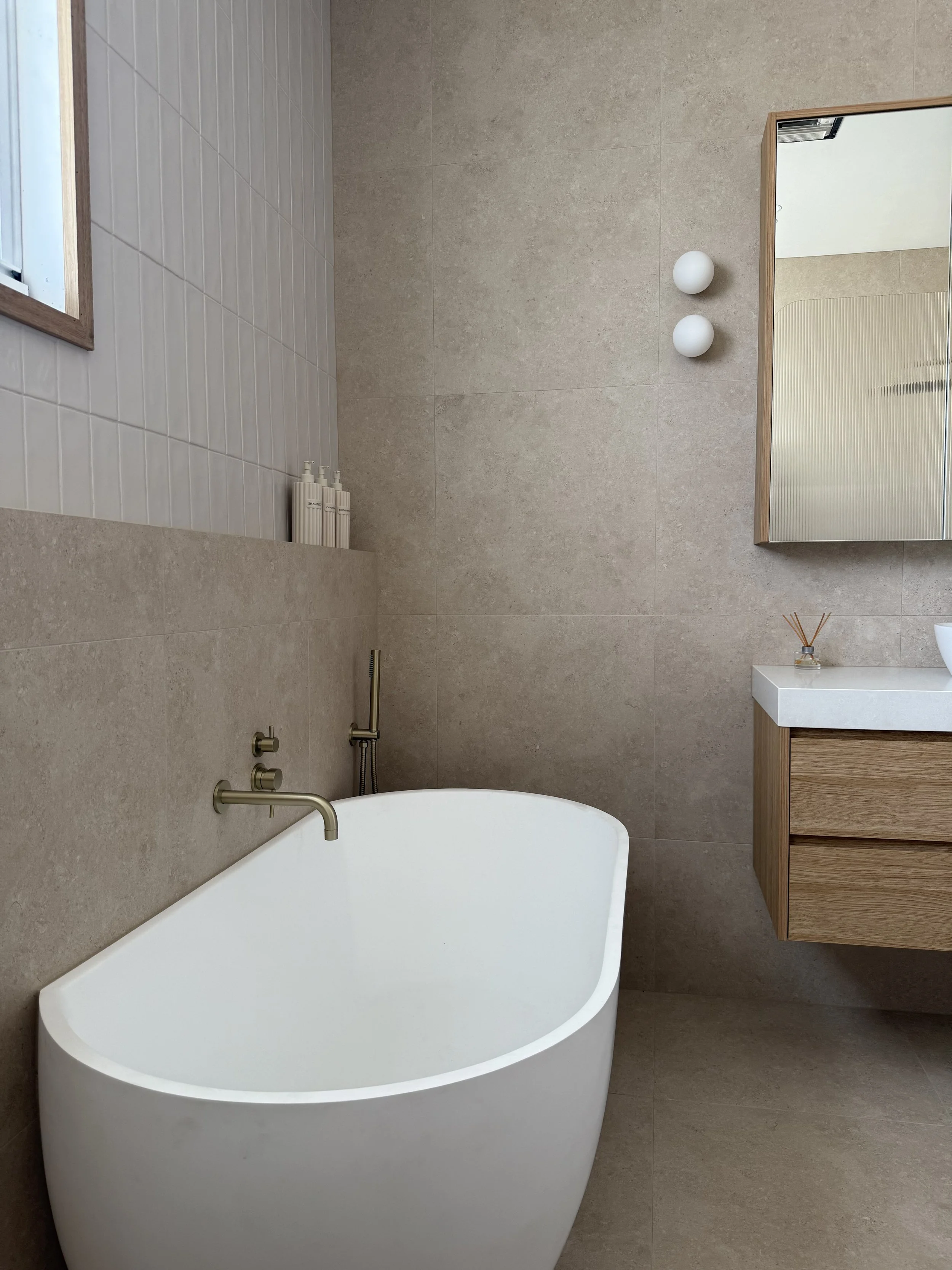 Modern bathroom with a freestanding white bathtub, a wall-mounted faucet, beige tiled walls, a small wooden vanity with a white countertop, a mirror, and decorative items.