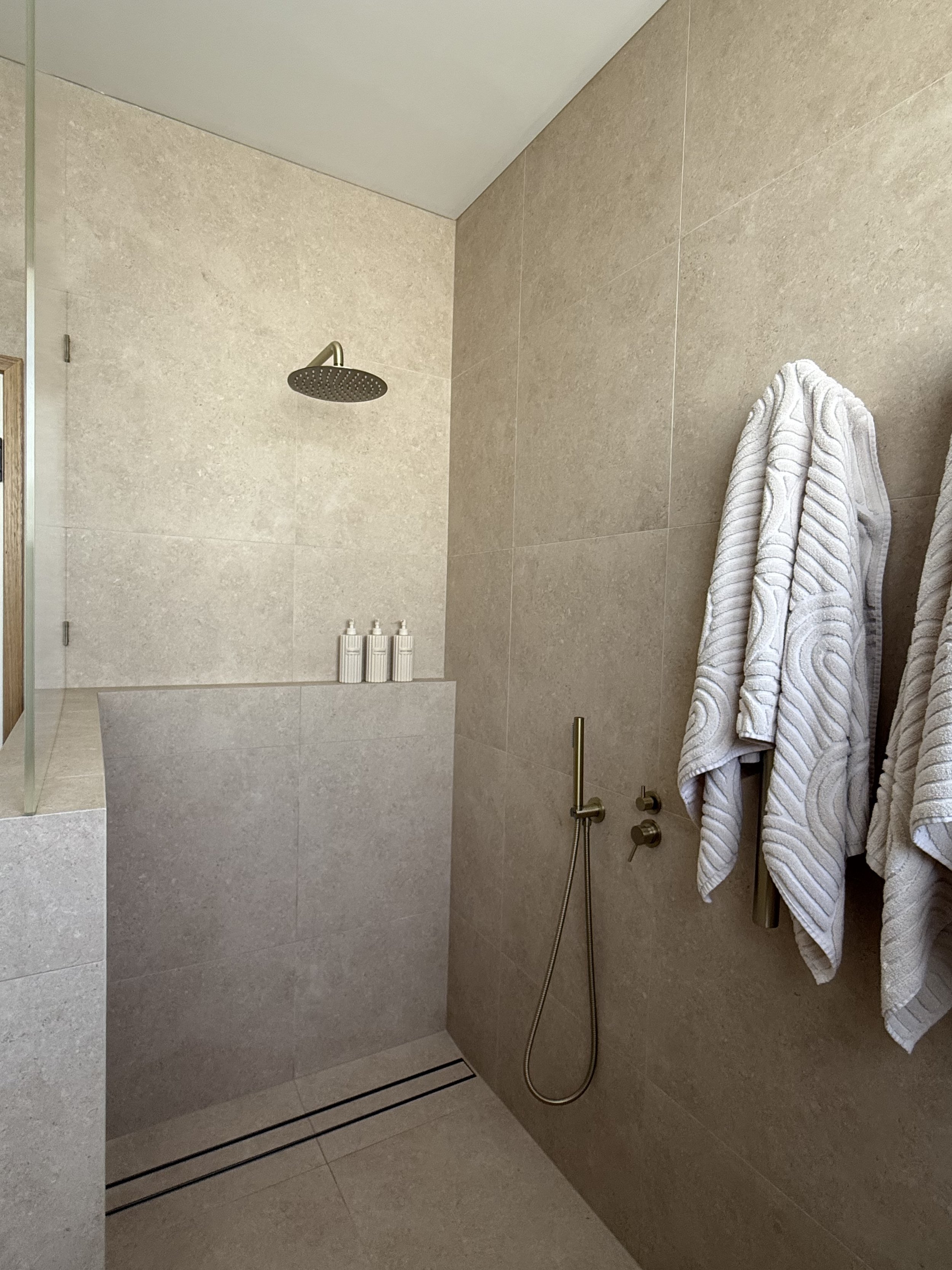 Minimalist shower area with a rain showerhead, beige tiled walls, a handheld shower, and white textured bathrobes hanging on hooks.