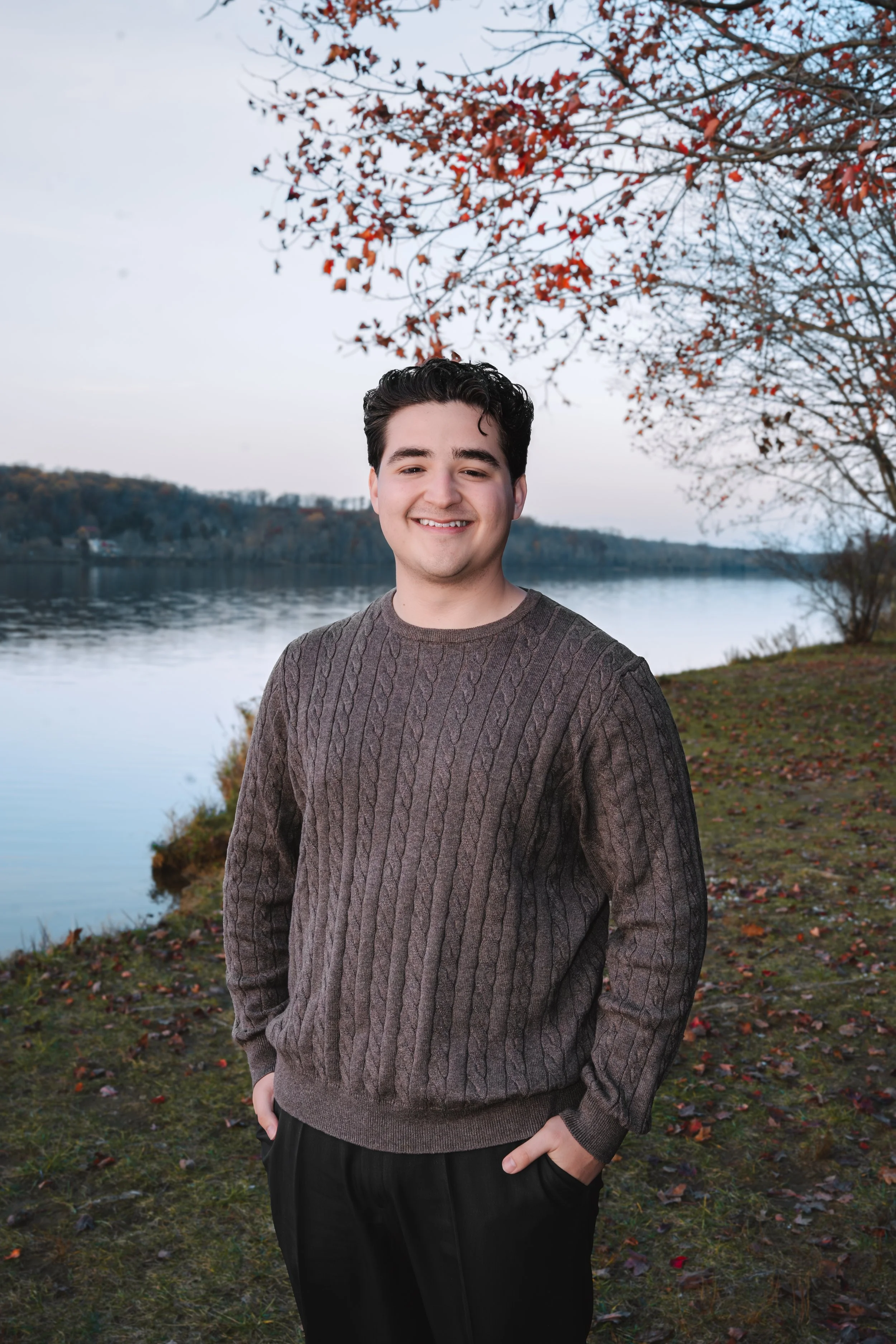 A young man smiling outdoors in autumn, standing near a calm body of water with trees displaying fall foliage in the background.