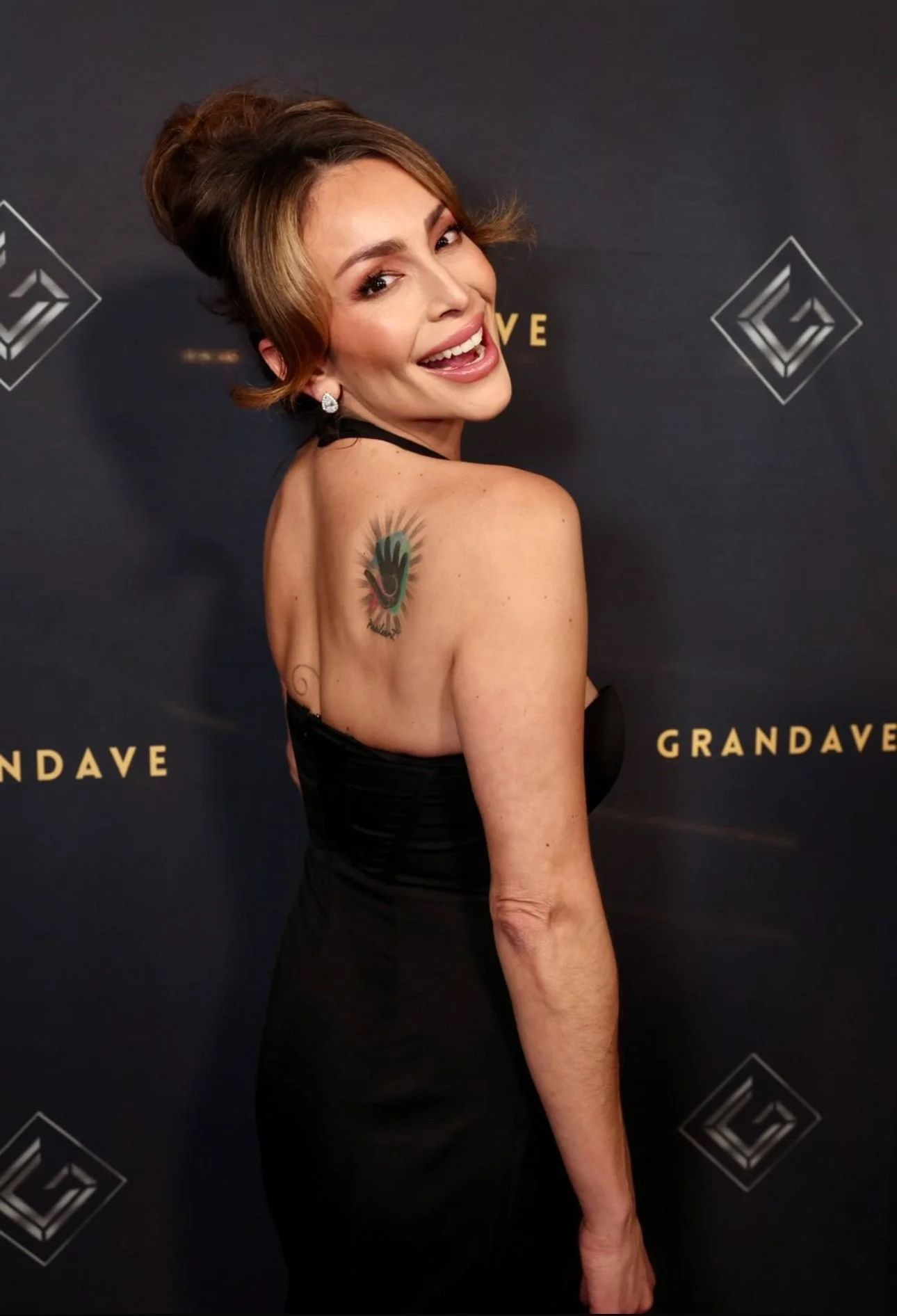A woman with short brown hair styled up, wearing a black strapless dress, smiling and looking over her shoulder, showing a colorful tattoo of a smiling face on her shoulder against a black background with gold text and logos.