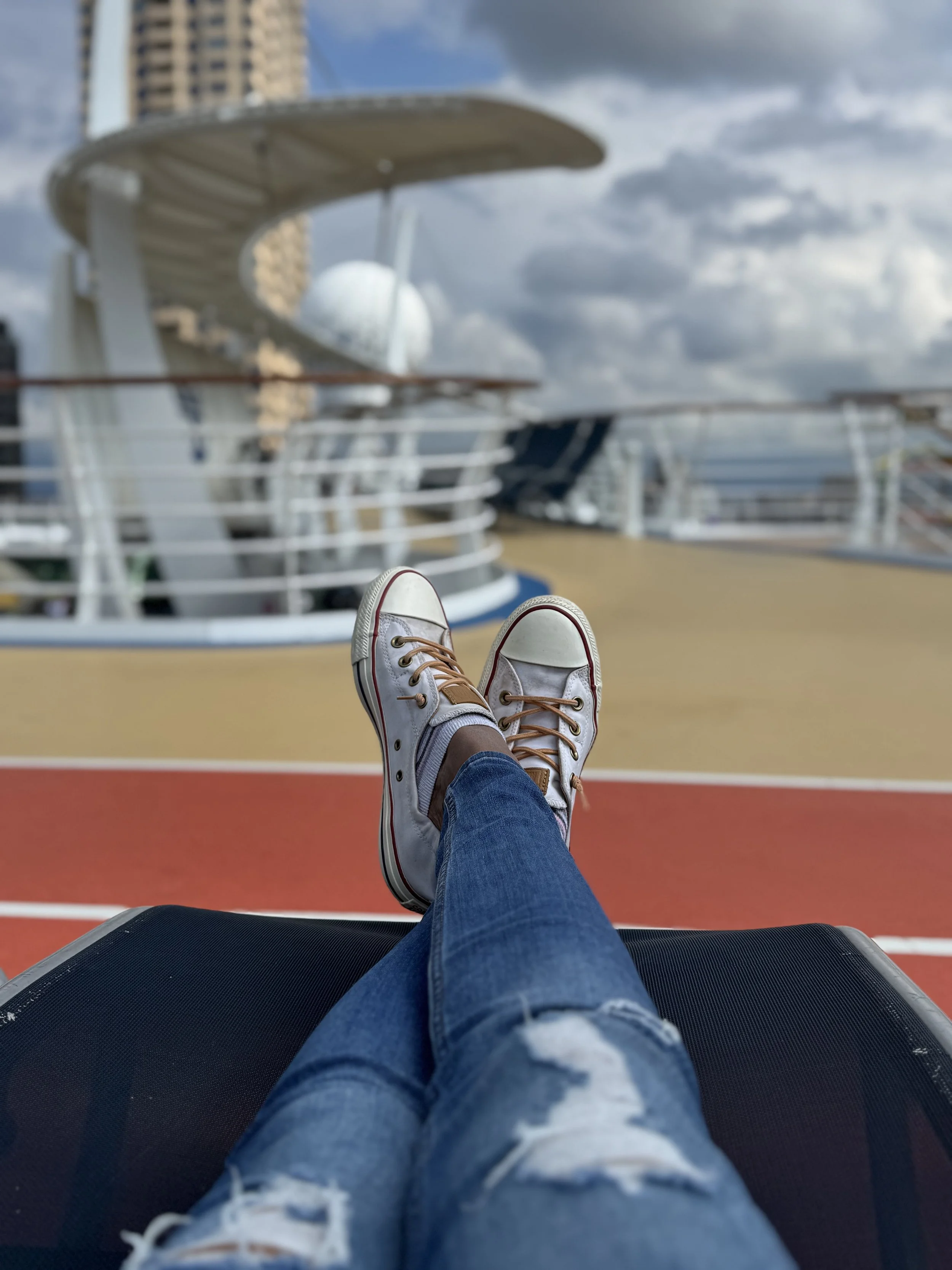 Person lounging on a deck chair with legs crossed, wearing white sneakers and ripped jeans, overlooking the cruise ship deck with a city skyline and cloudy sky in the background.