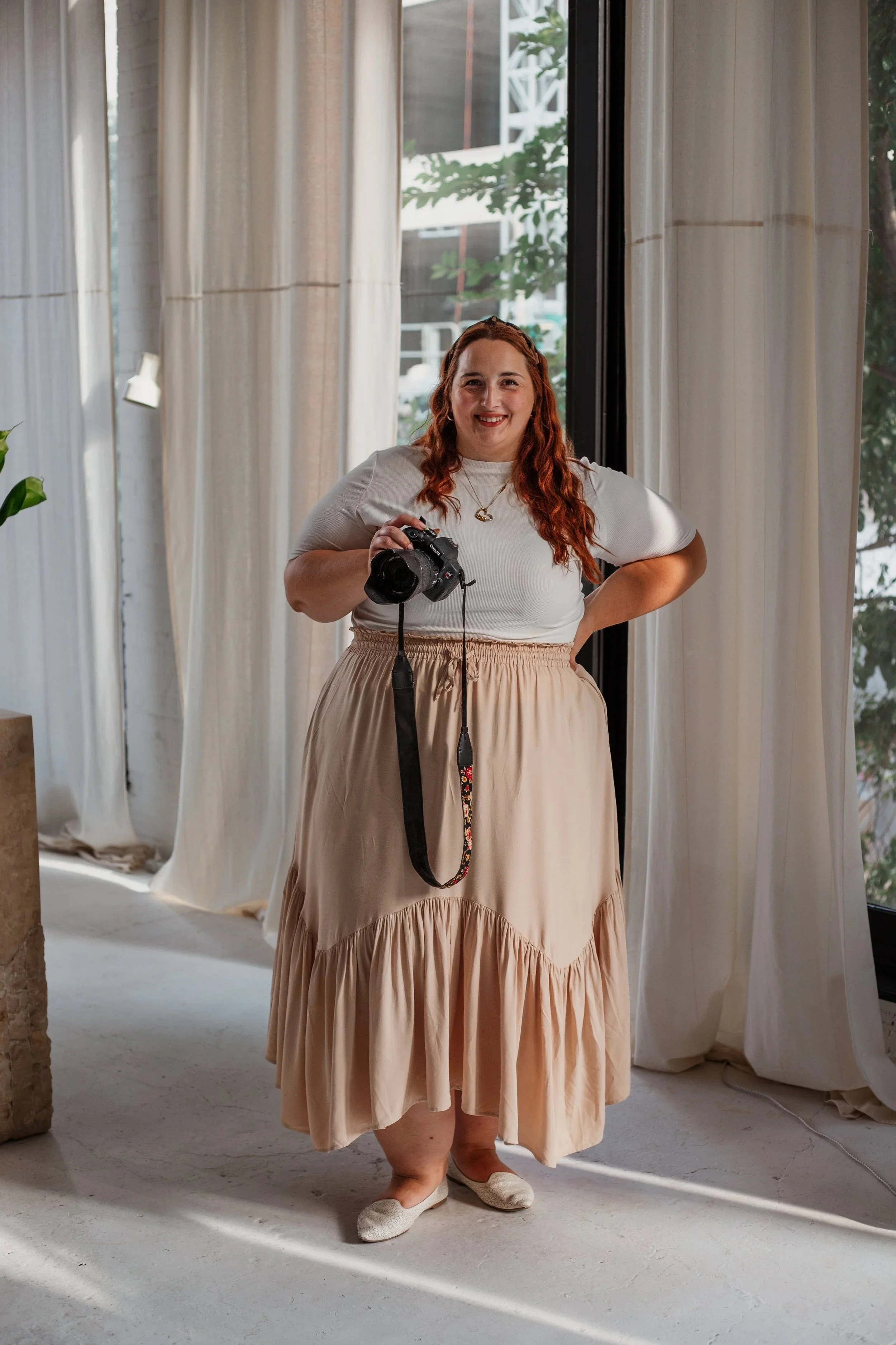 A woman with long red wavy hair, wearing a white top and beige skirt, is standing indoors near a large window with white curtains, holding a camera and smiling at the camera.