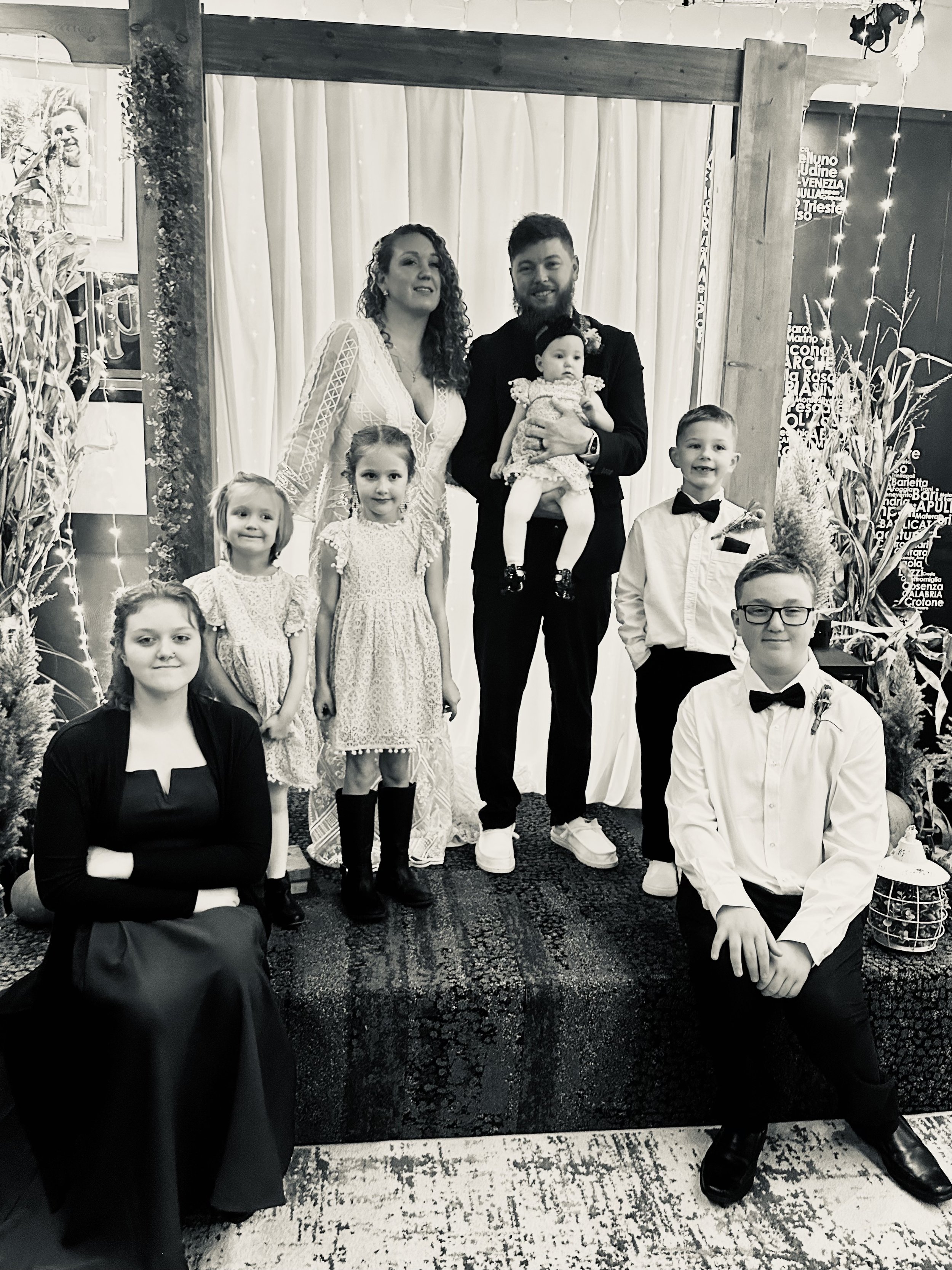 A family photo taken at a celebration, with adults and children dressed in formal attire, standing in front of a decorated backdrop with curtains and fairy lights.