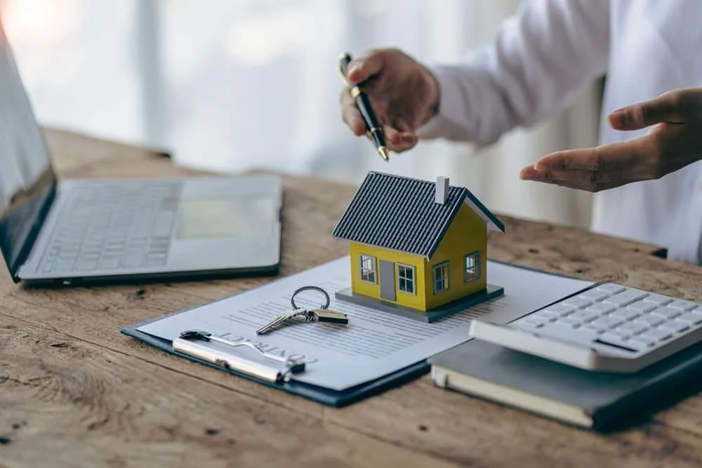 Person reviewing a small model house on a wooden table with a laptop, keyboard, clipboard with papers, and keys.