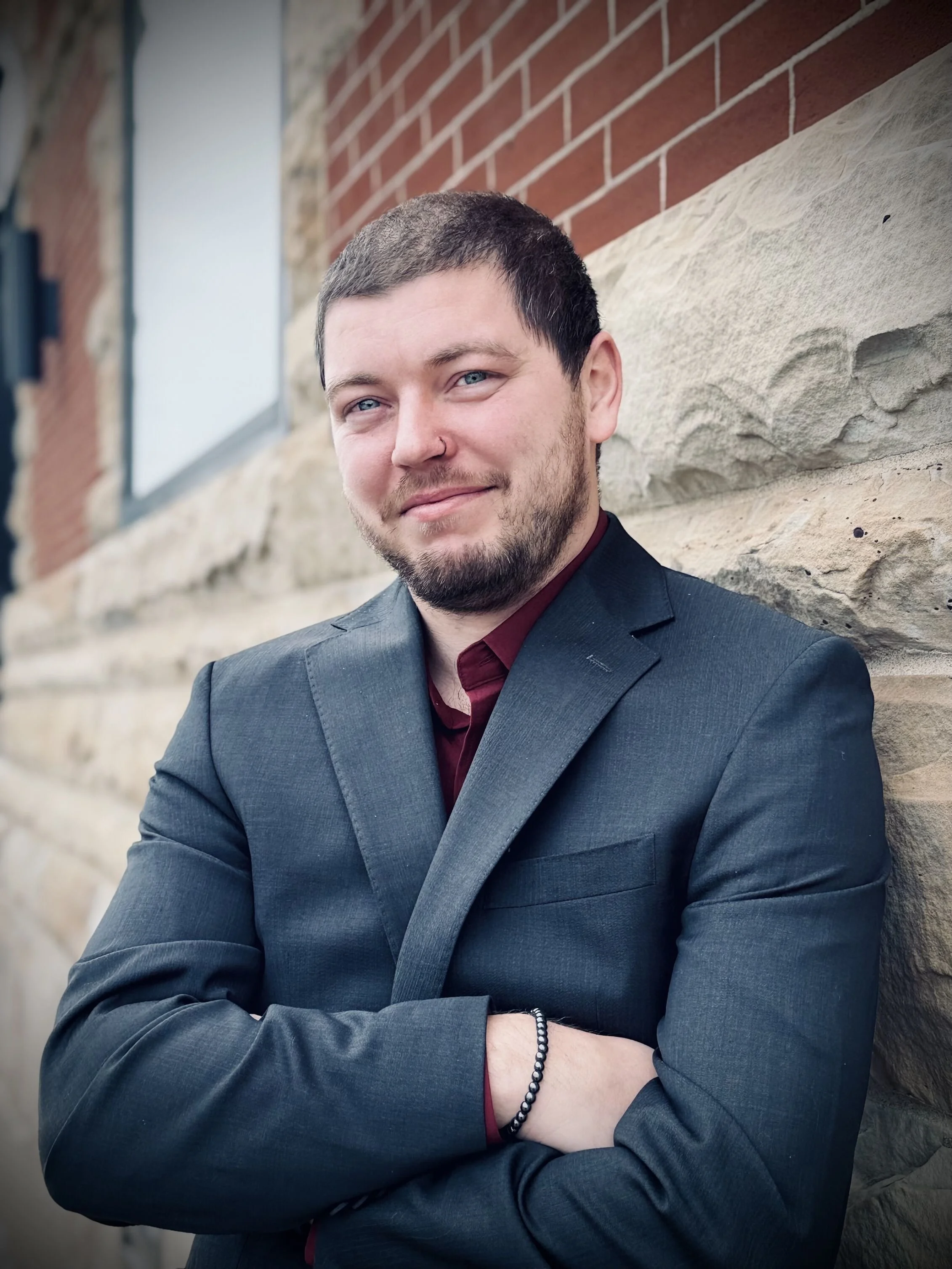 A young man with short dark hair and a beard, dressed in a dark gray suit jacket and a maroon shirt, standing outdoors against a brick and stone wall with his arms crossed and smiling.