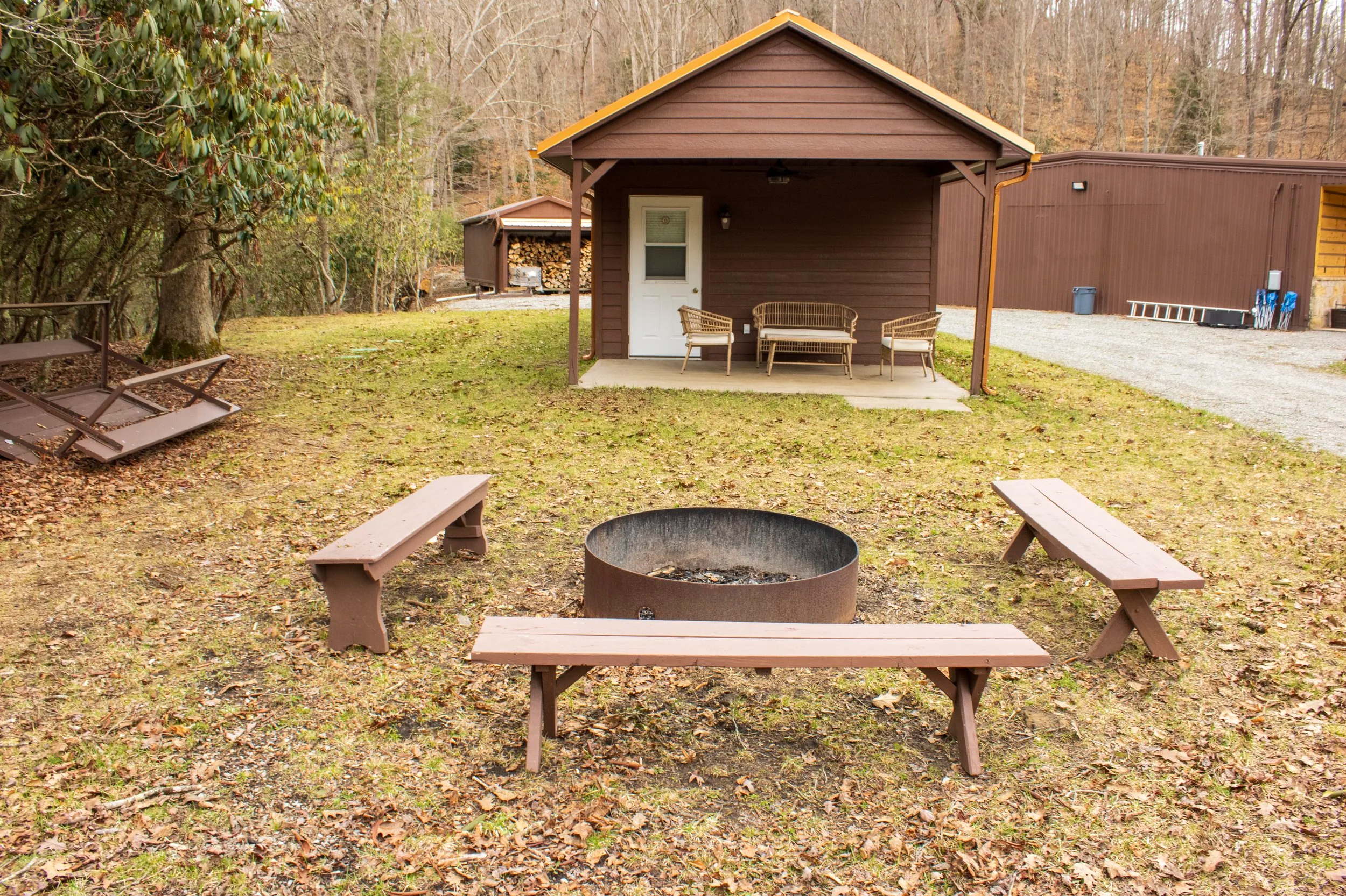 A backyard scene with a firepit surrounded by benches, a grass yard, a small brown house with a porch and outdoor furniture, a gravel pathway, and a larger building in the background, with trees and fallen leaves in autumn.