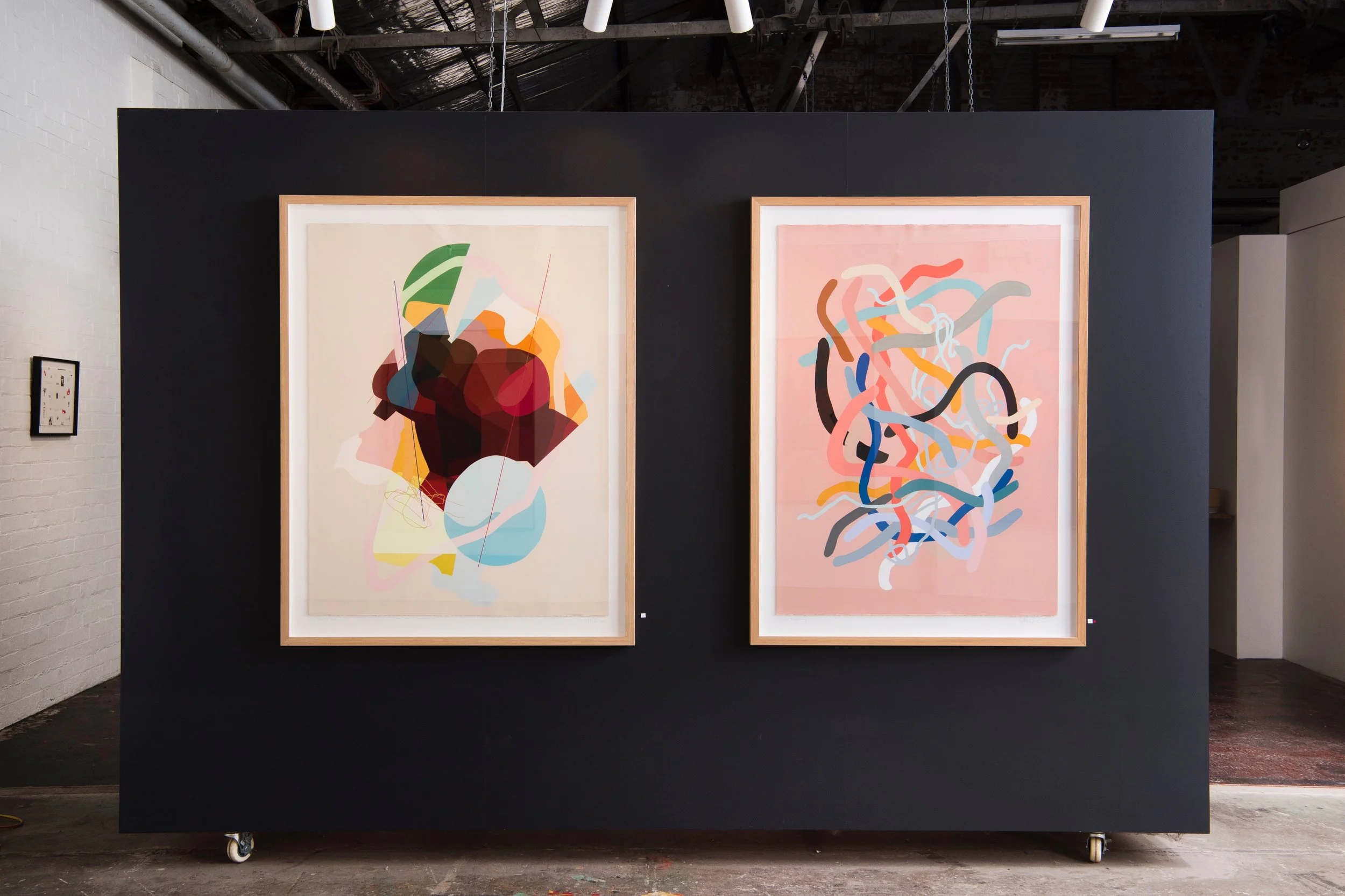 Two abstract paintings hanging on a dark gallery wall, each framed in light wood, with a smaller artwork on the left wall.