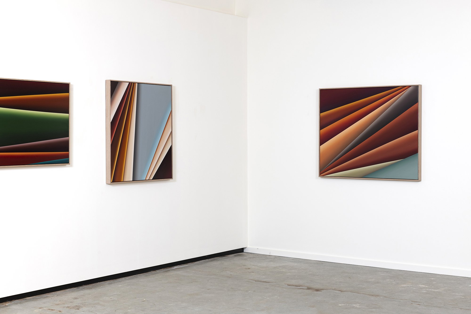 Three abstract colorful framed artworks hanging on a white gallery wall with a concrete floor.