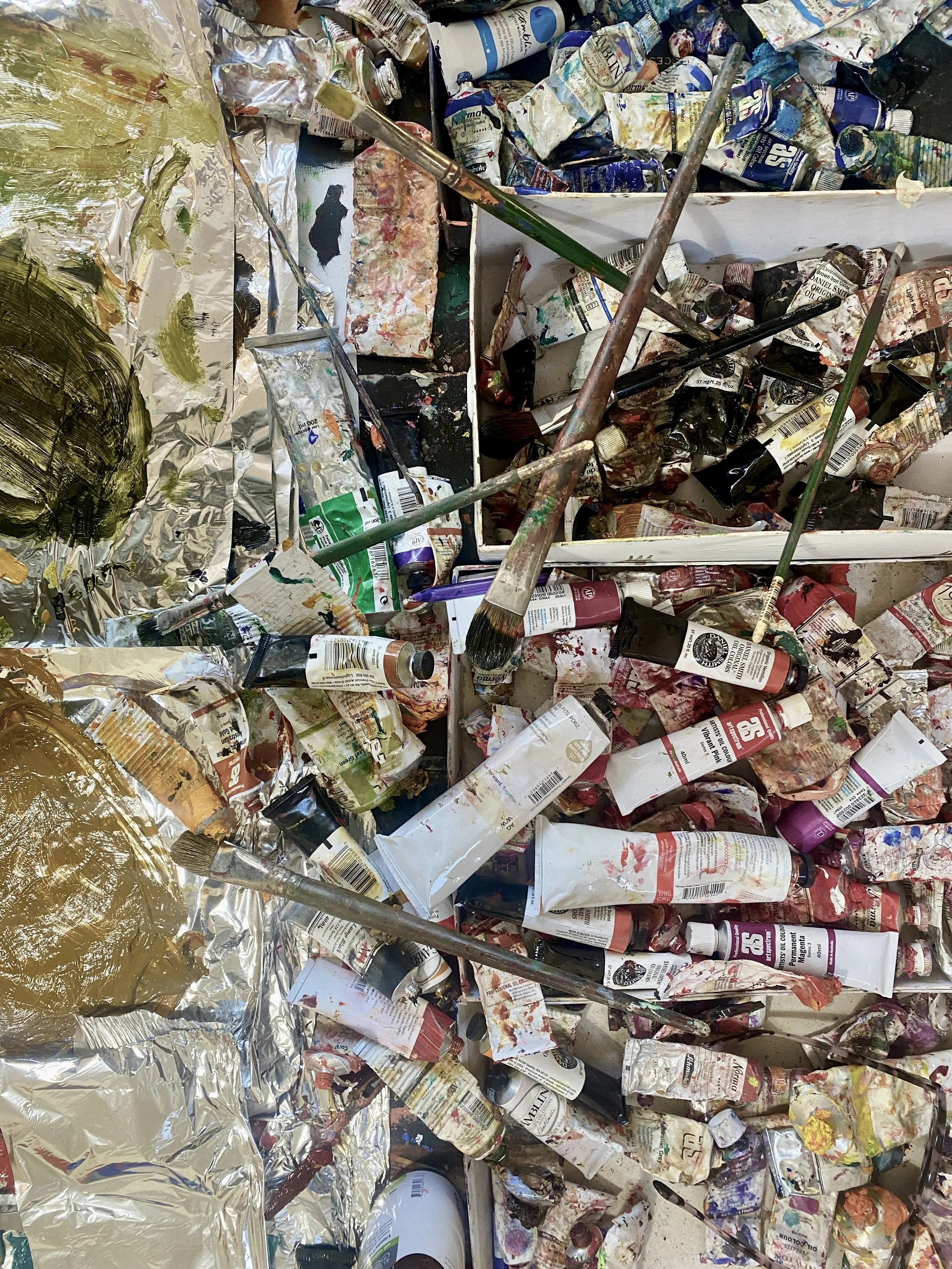 A cluttered art station with numerous paint tubes, brushes, and supplies scattered around, some covered in dried paint, with aluminum foil and plastic wrapping also visible.