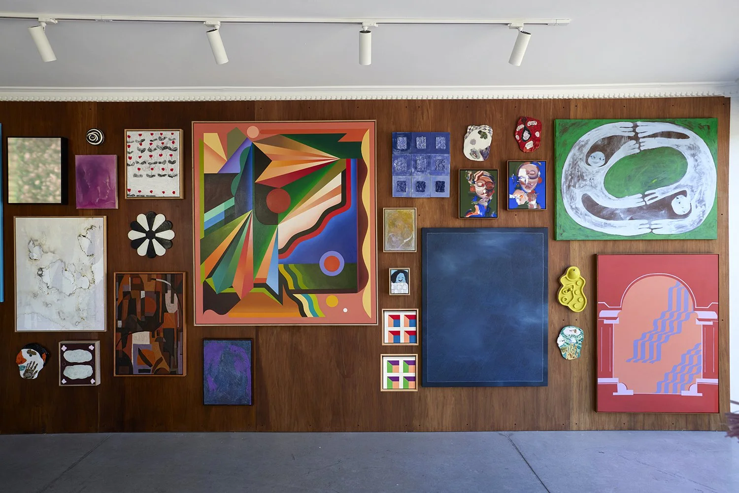 An art gallery wall with a variety of colorful paintings and mixed media art pieces displayed on a wooden panel background.