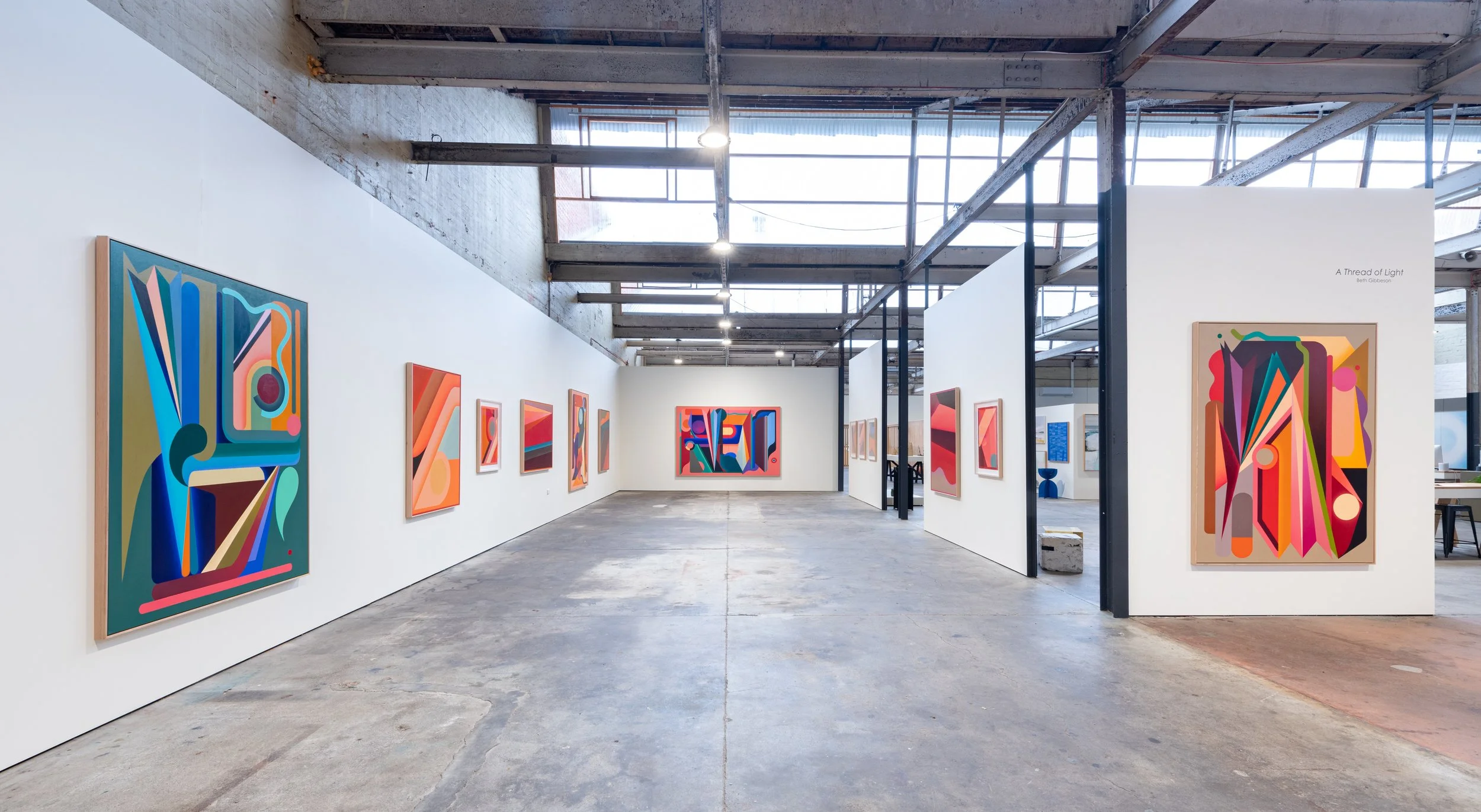 An art gallery with abstract colorful paintings displayed on white walls in a spacious industrial-style interior.