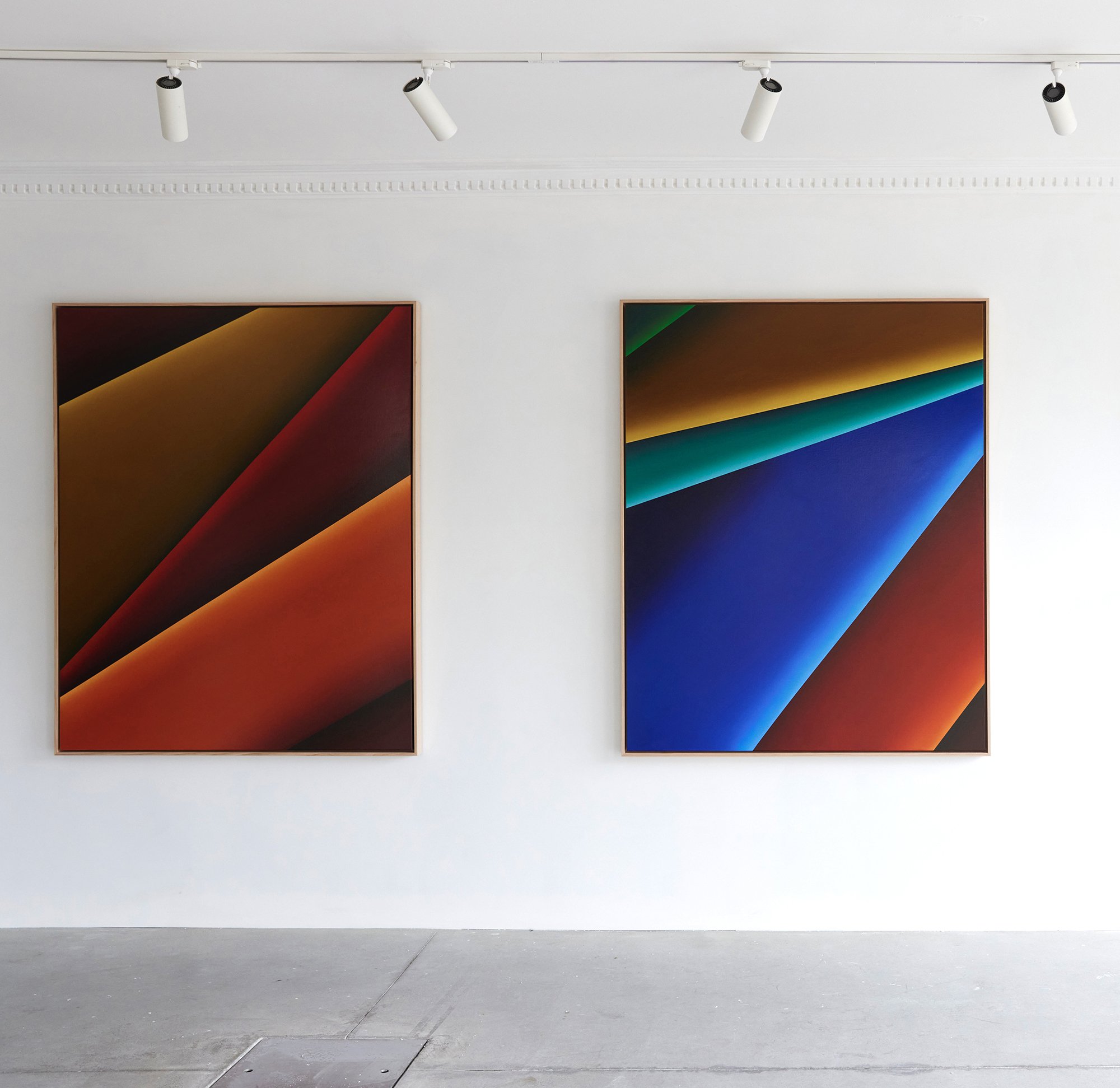 Two abstract colorful paintings hanging on a white gallery wall, lit by ceiling spotlights.