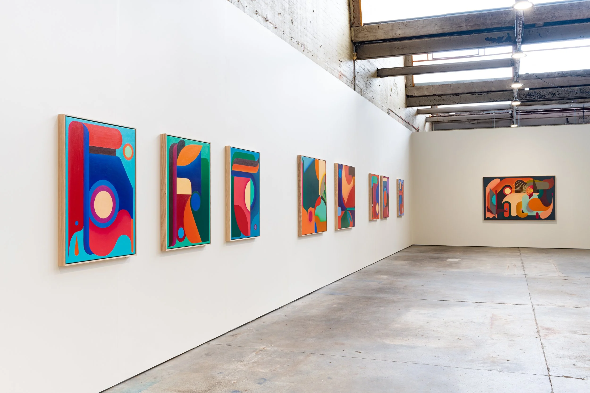 An art gallery with eight colorful abstract paintings hanging on a white wall. The gallery has a concrete floor and skylights with visible wooden beams on the ceiling.