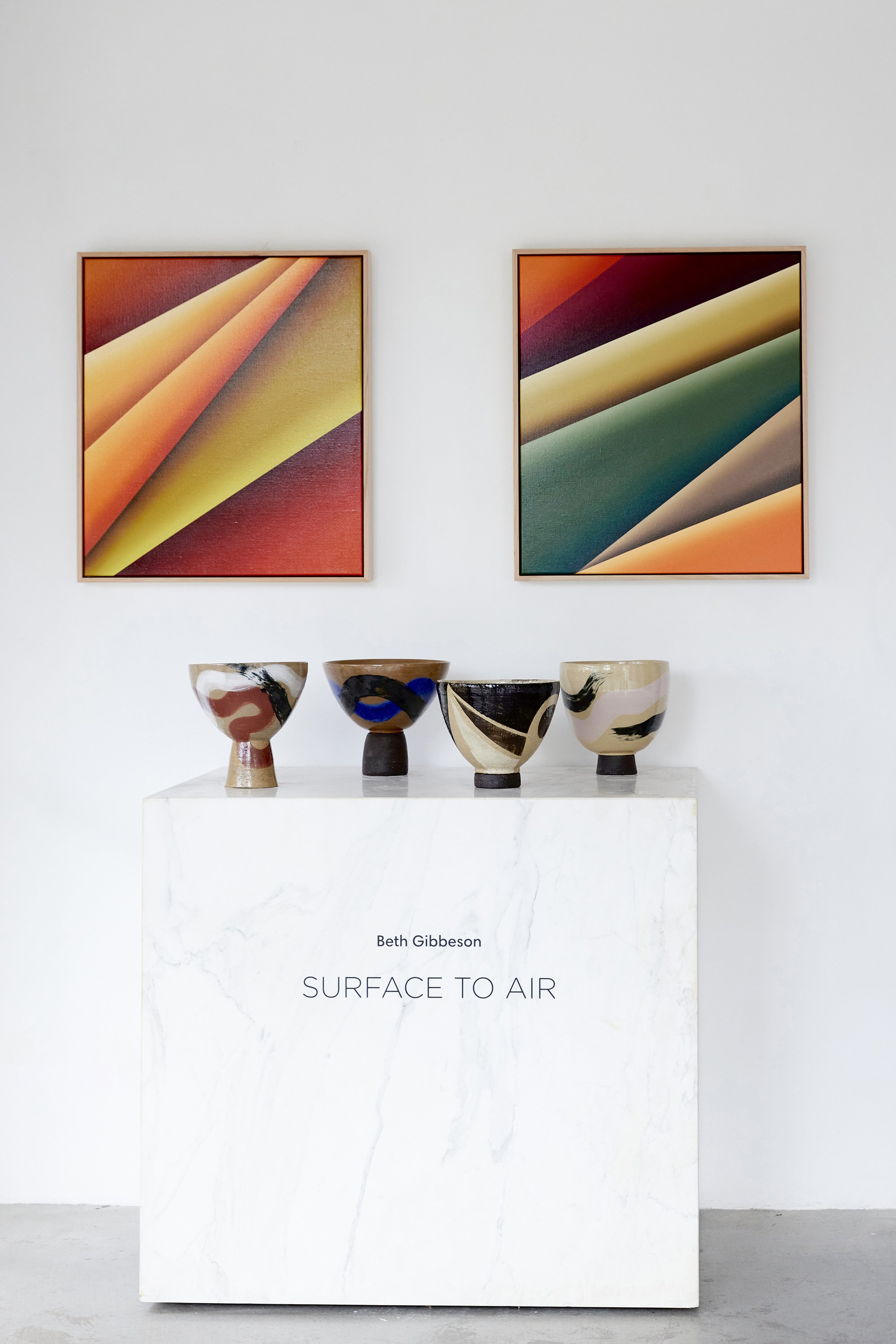 Art exhibit display with four ceramic bowls on a white pedestal in front of two abstract colorful paintings on a white wall.