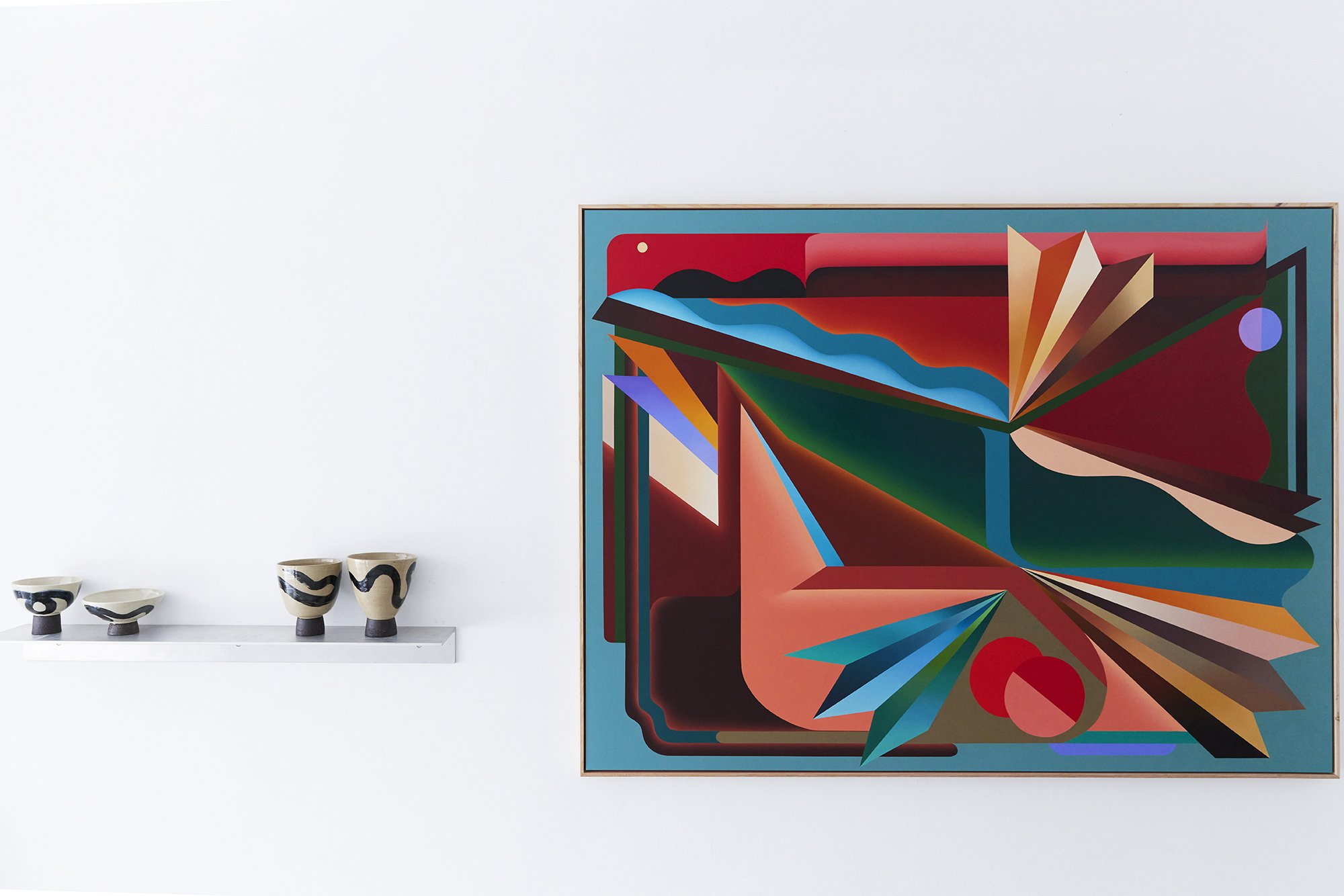 An abstract geometric painting with vibrant colors on a white wall, with four black and white ceramic cups on a shelf below.