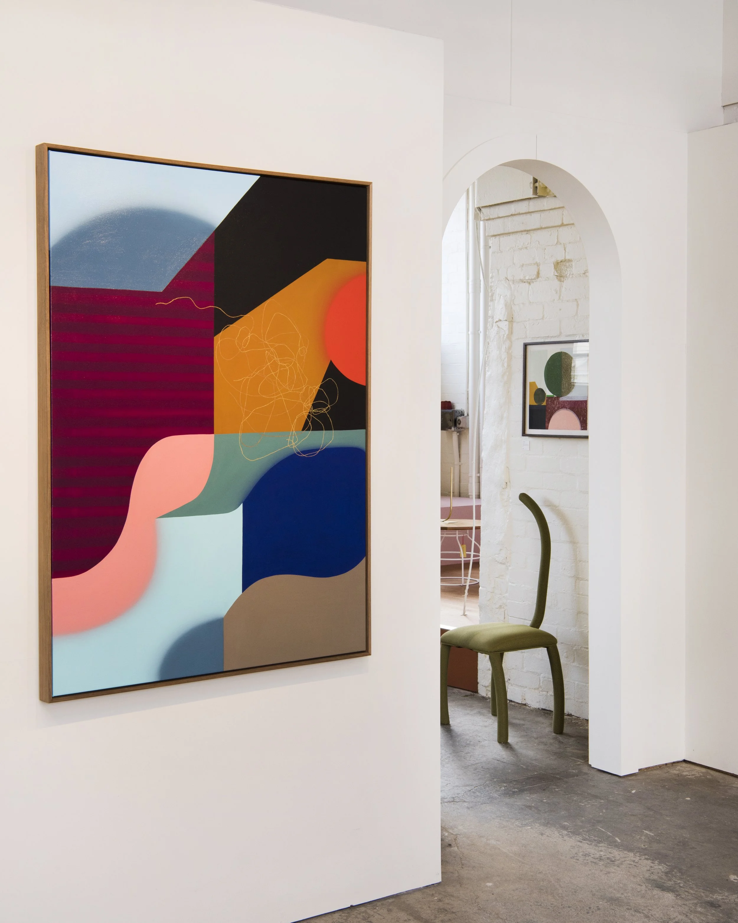 Abstract colorful artwork on a white gallery wall with a chair visible through an arched doorway.