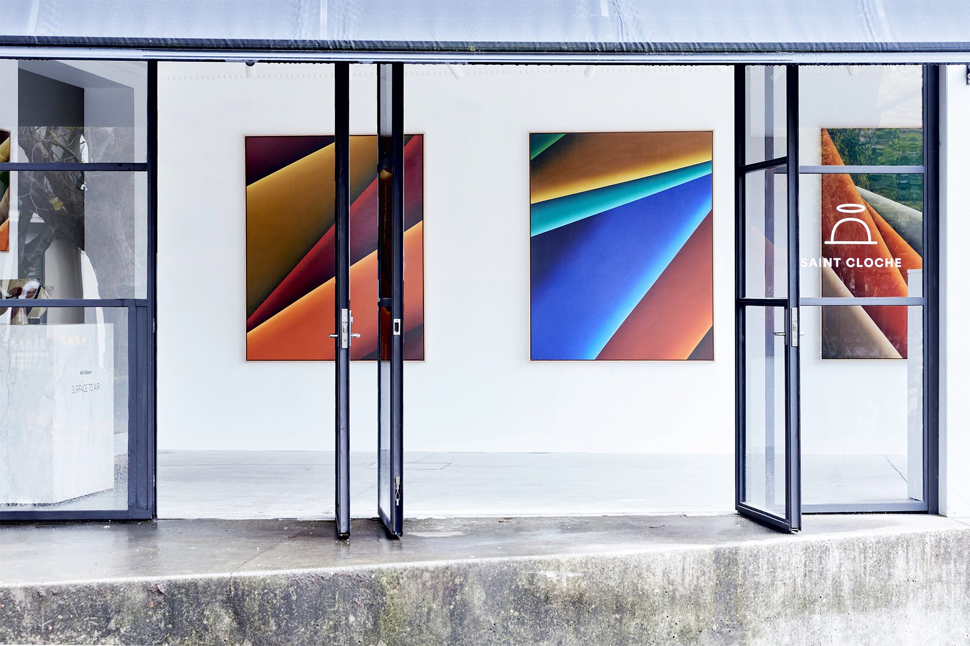 View of an art gallery with three colorful abstract paintings on white walls, glass doors open to the outside, and the text 'Saint Cloche' on the right door.
