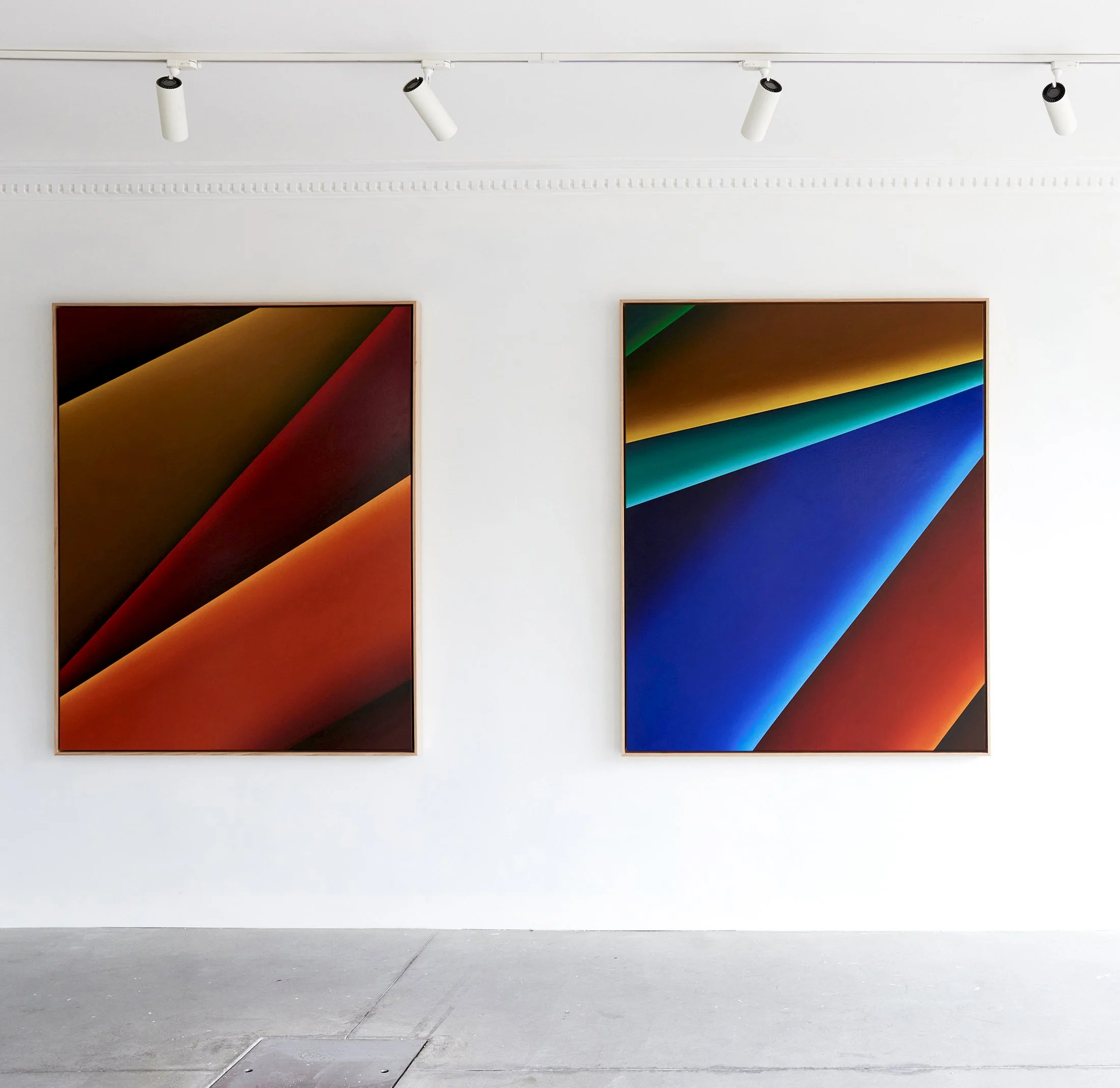 Two colorful abstract paintings with diagonal lines, hung on a white gallery wall, illuminated by track lighting.