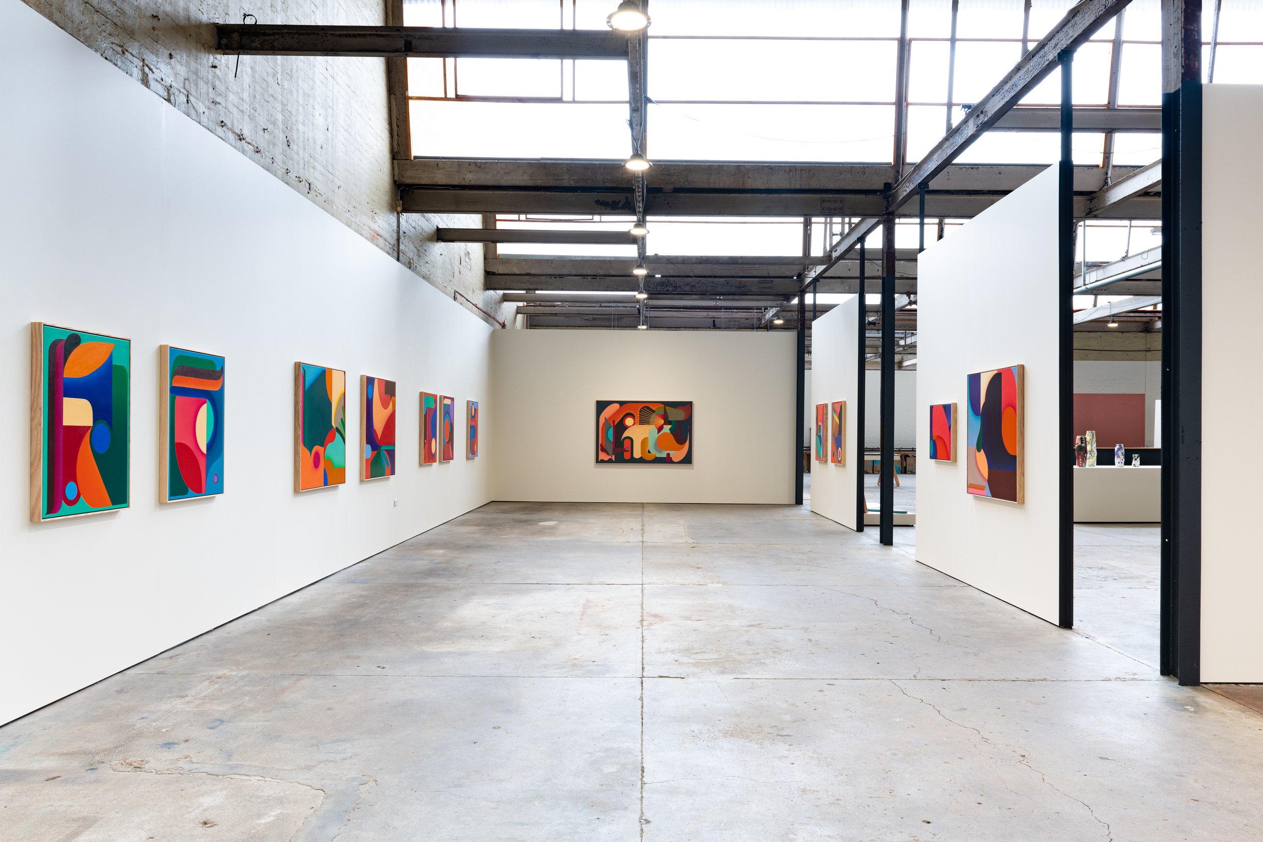 Inside an art gallery with white walls displaying colorful abstract paintings. The space has a high ceiling with exposed beams and skylights, concrete floors, and partitions creating separate sections.