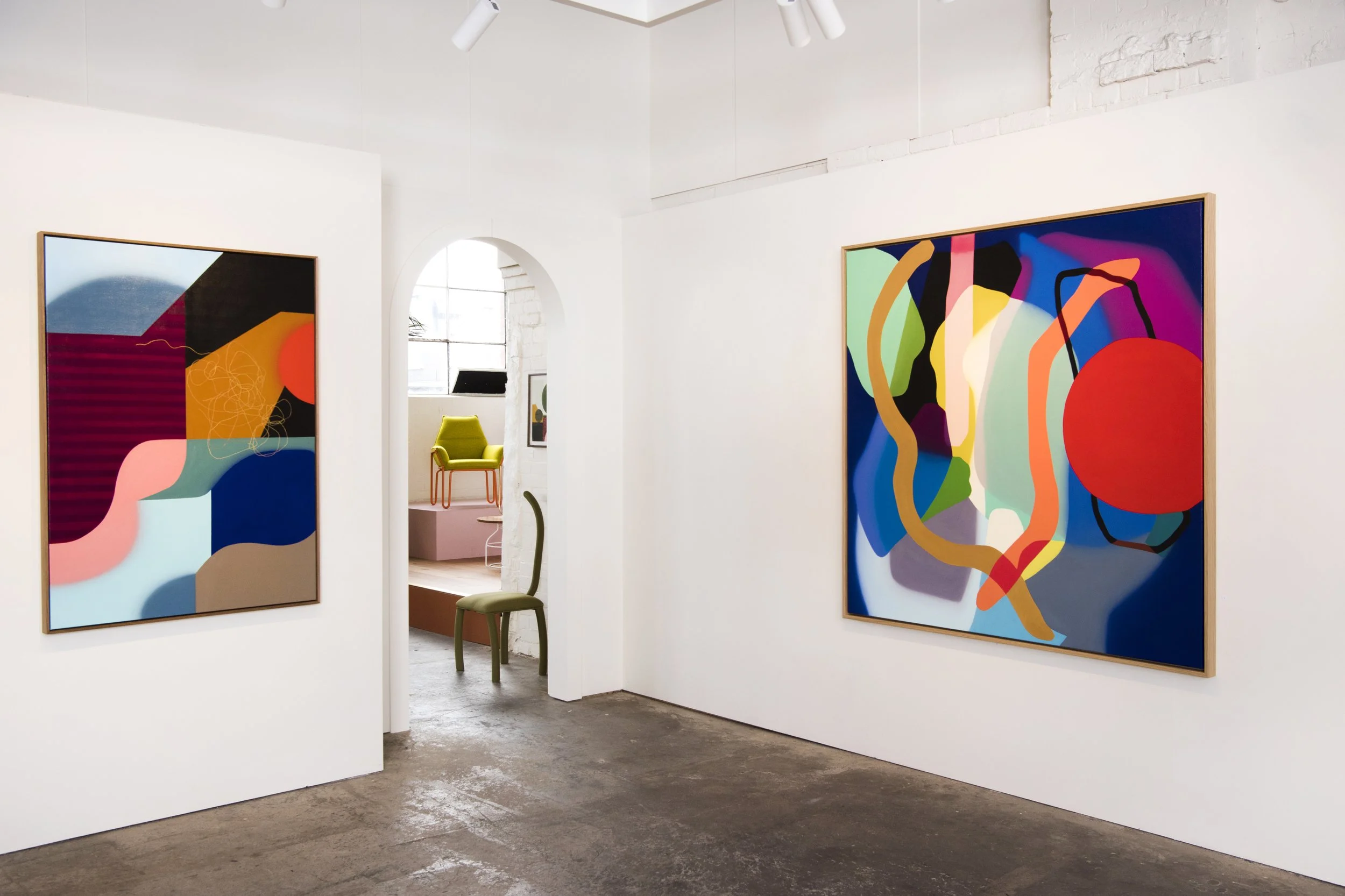 An art gallery with two large colorful abstract paintings on white walls, a green chair in the foreground, and a small room with more artwork and a green chair visible through an arched doorway.