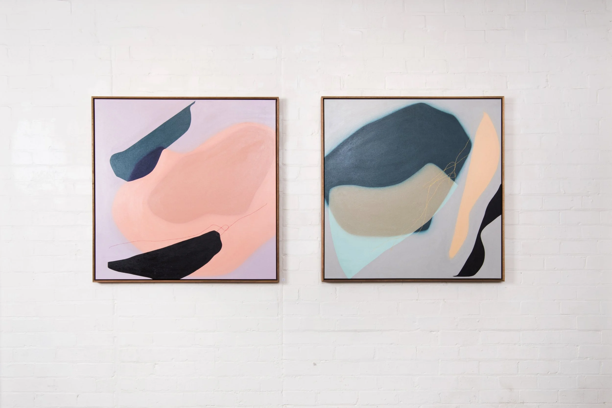 Two abstract paintings with organic shapes in pastel and darker colors, framed and hanging on a white brick wall.