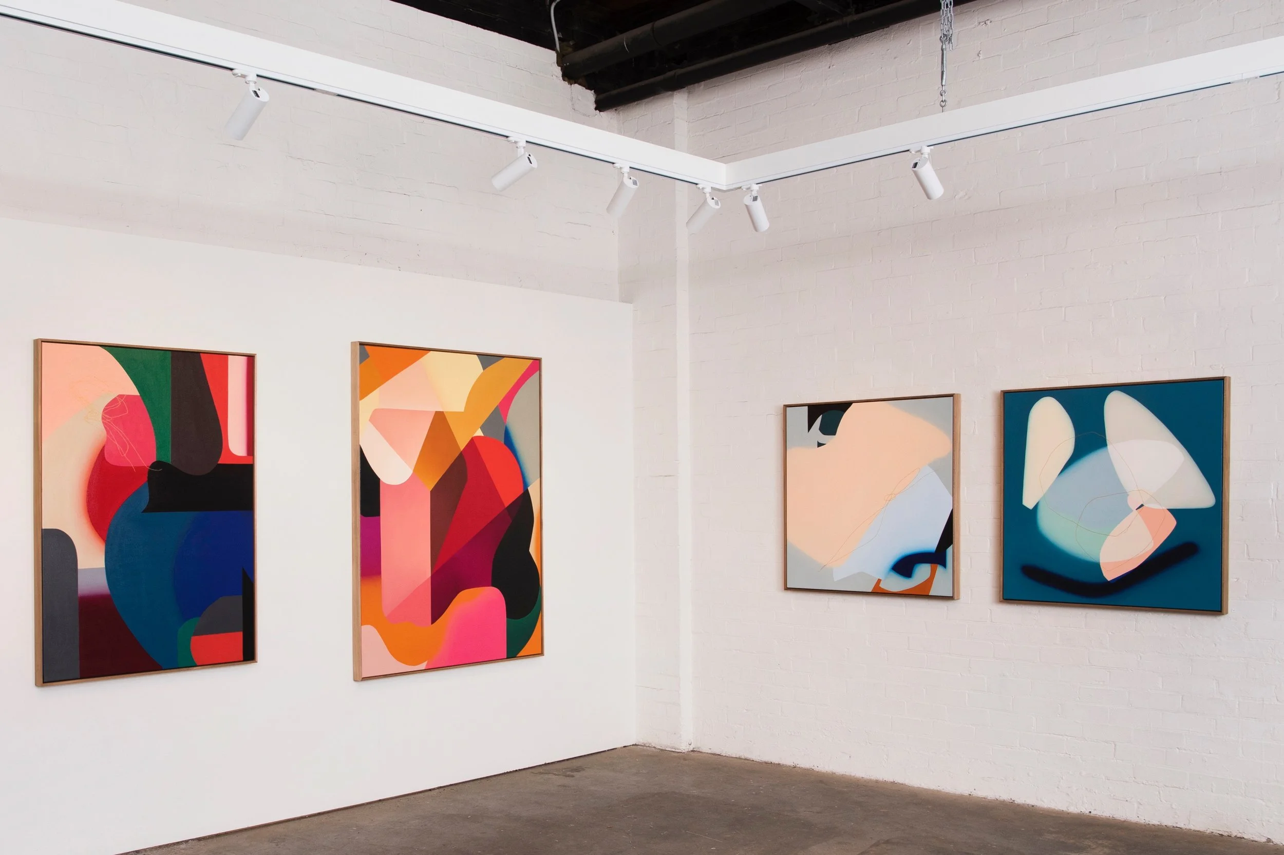 An art gallery with four abstract paintings on white brick walls, illuminated by ceiling track lighting.